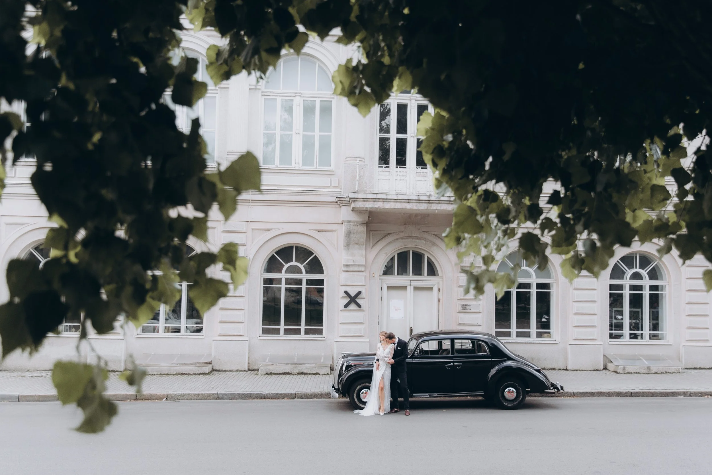 A couple in wedding attire stands kissing in front of a vintage black car on a city street, with white buildings and large arched windows in the background, framed by green tree leaves at the top.