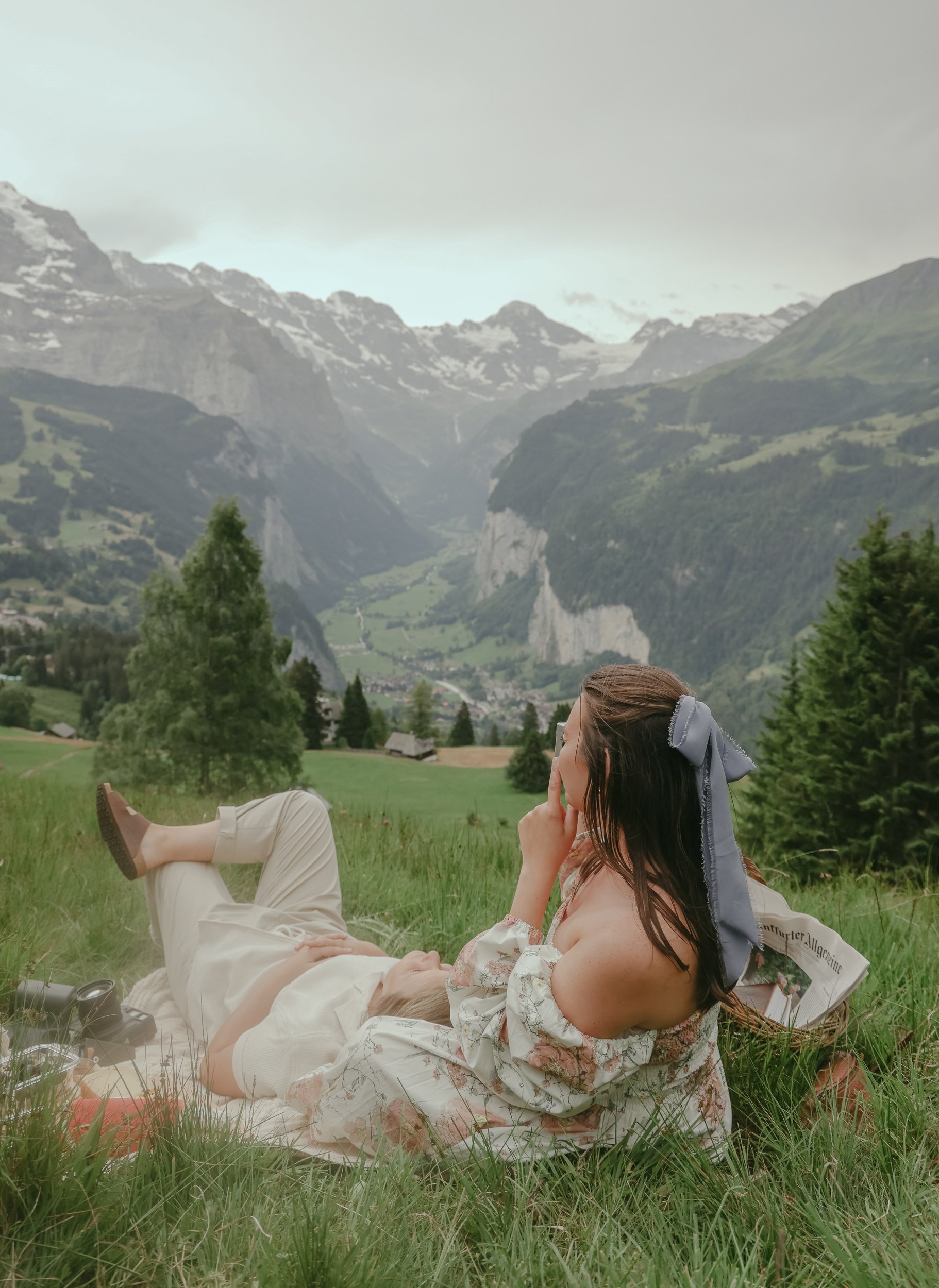 A woman with long brown hair tied with a blue ribbon and wearing an off-shoulder dress is laying on the grass next to a person lying on their back. They are in a green field with trees and mountains in the background, under a cloudy sky.