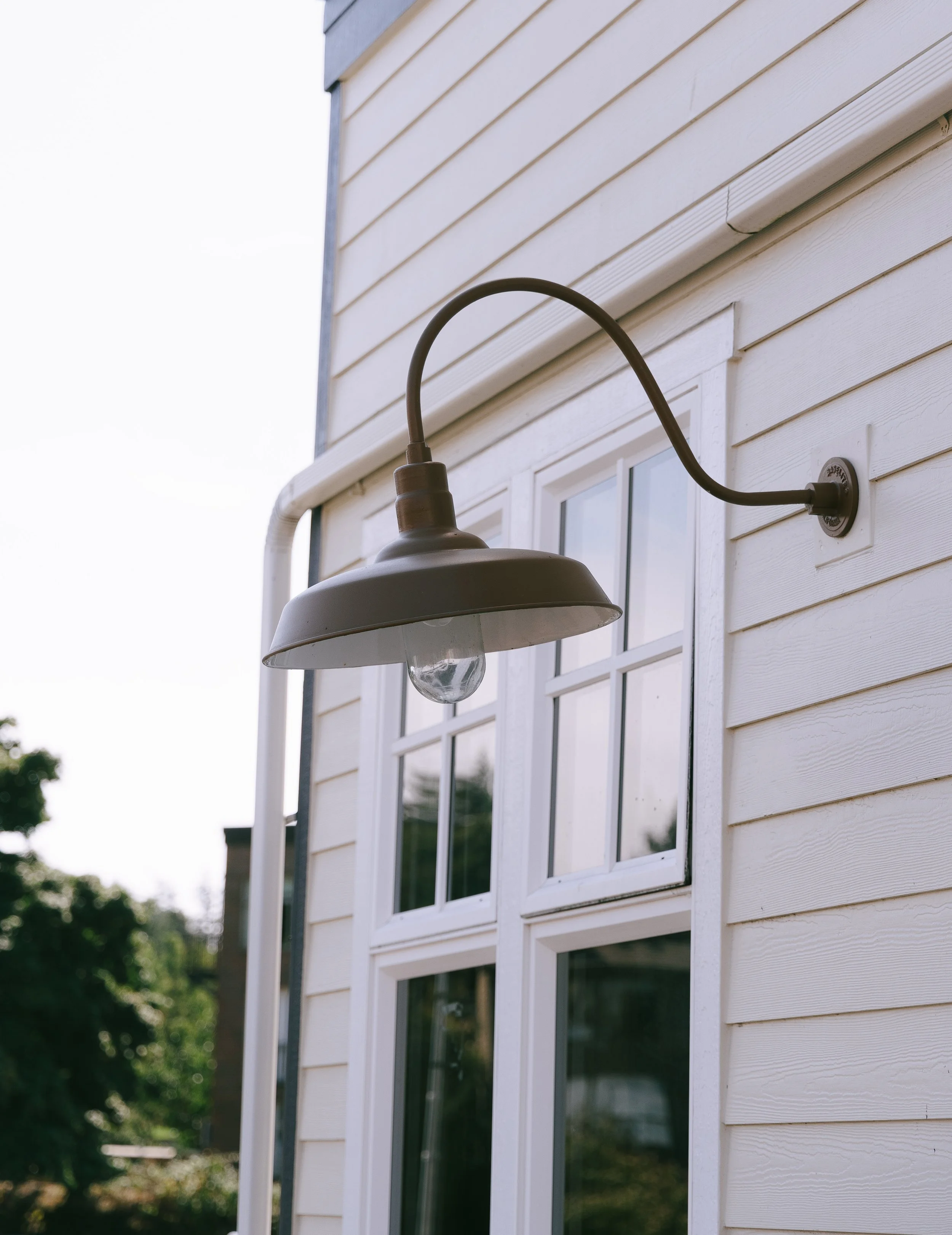 Exterior of a house with a white wooden wall and a window, featuring an outdoor wall-mounted light fixture with a curved arm and a clear glass bulb.