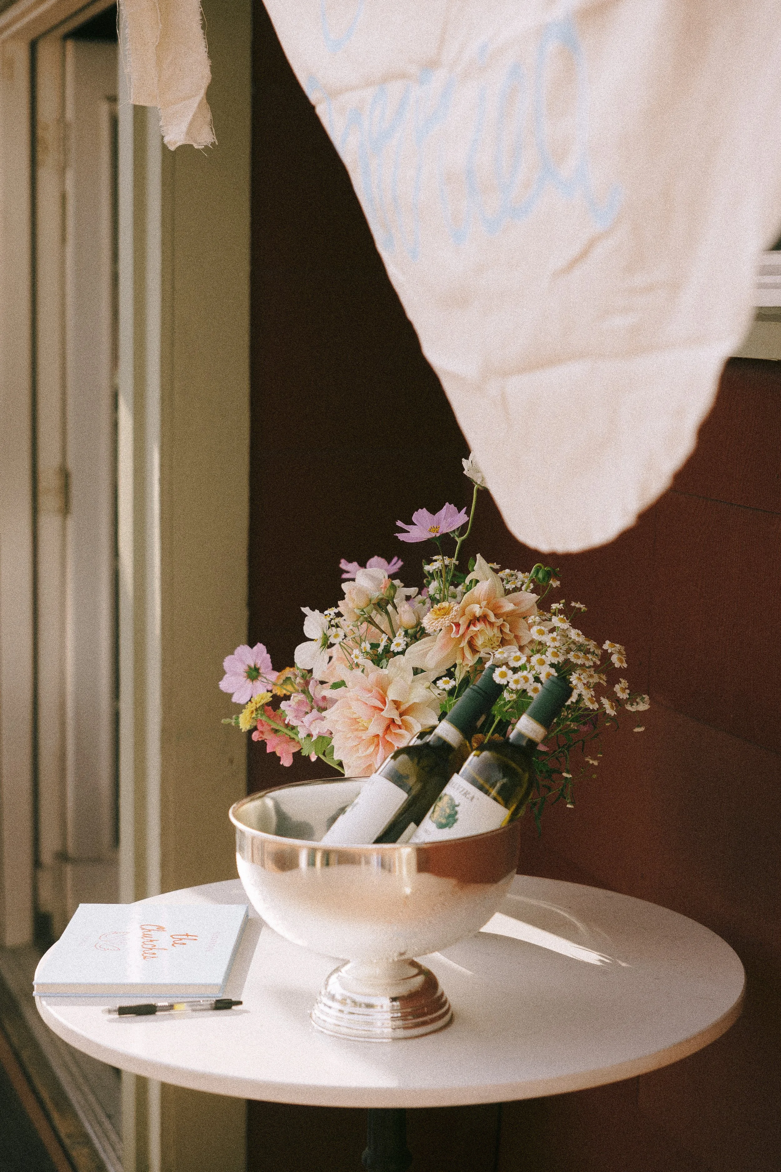 A white tabletop with a silver bowl containing two bottles of wine, a bouquet of pink and white flowers, and a small white book with orange text and a black pen on top. Part of a paper bag is hanging above.