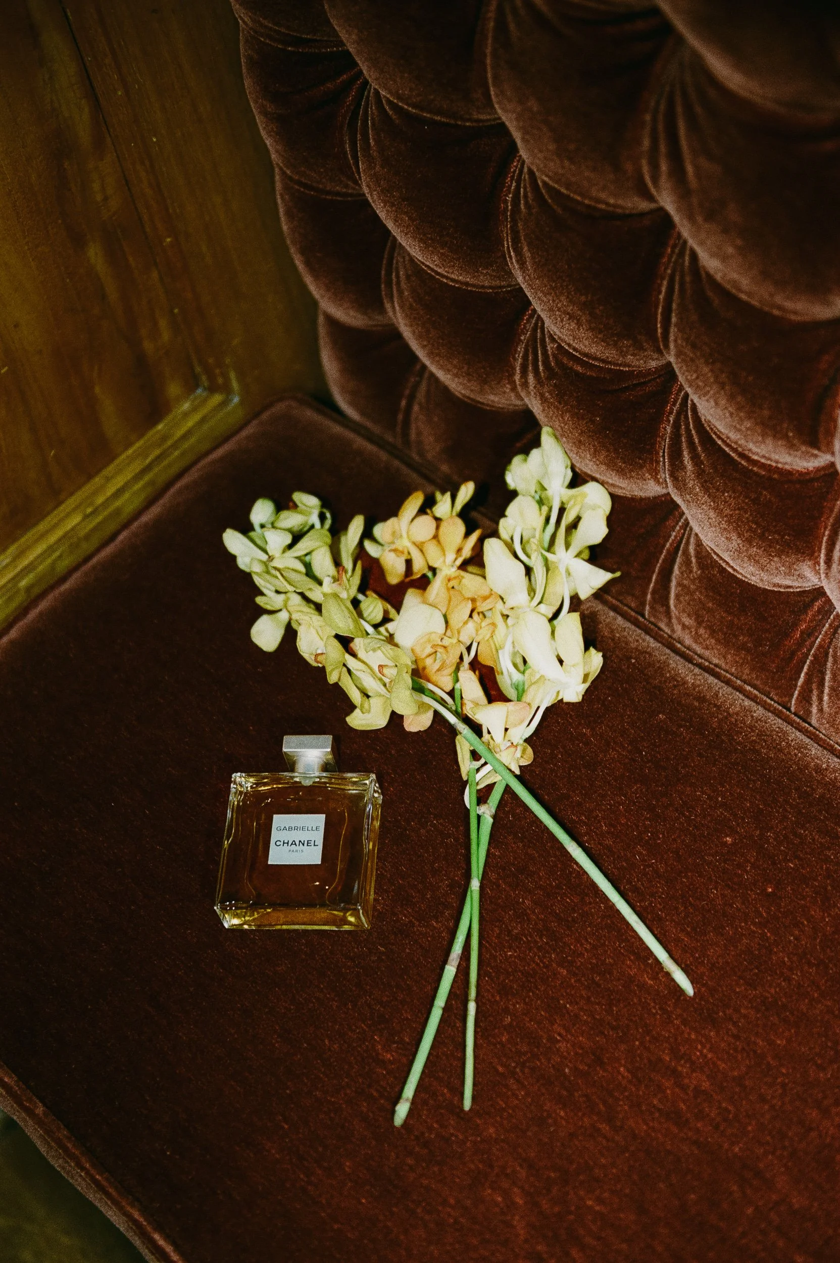 A bottle of Chanel Gabrielle perfume and a small bouquet of wilted white flowers on a vintage brown velvet chair.