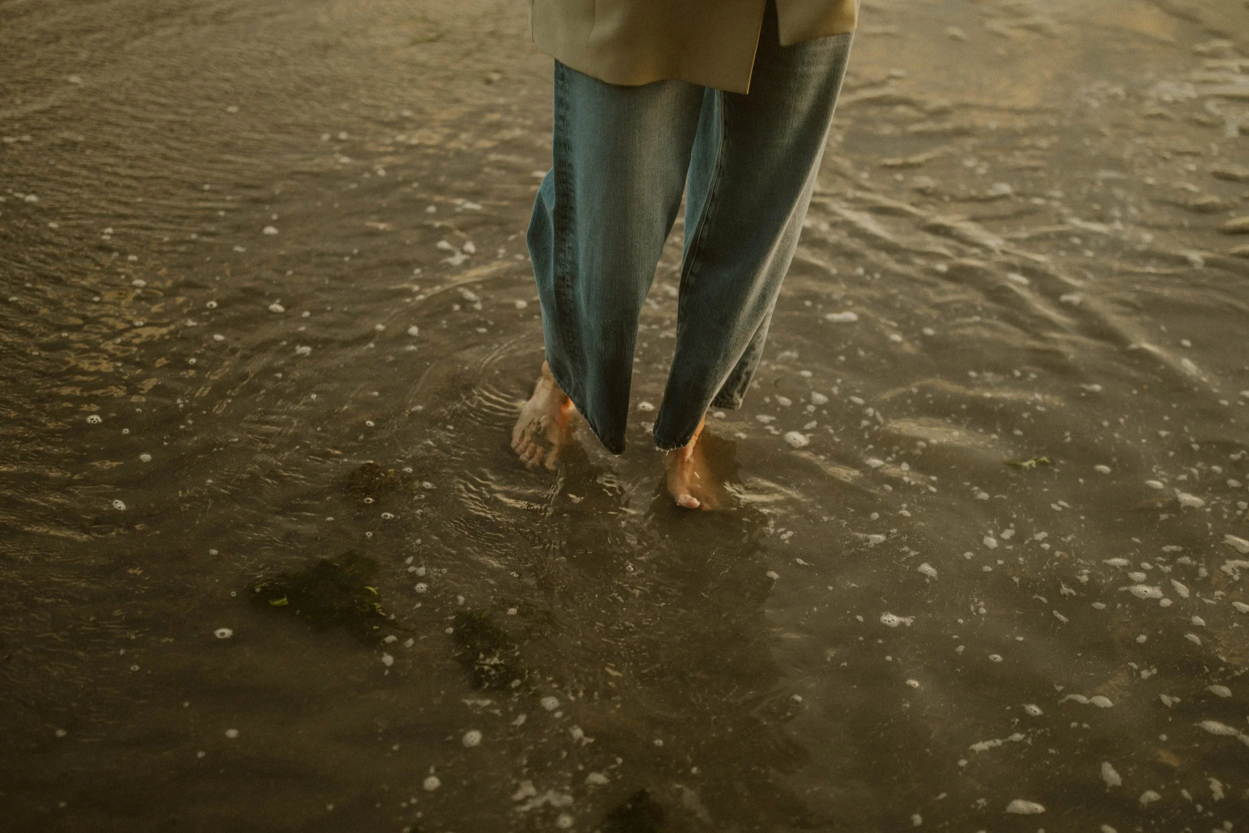 Person walking barefoot in shallow water wearing jeans and a beige top.