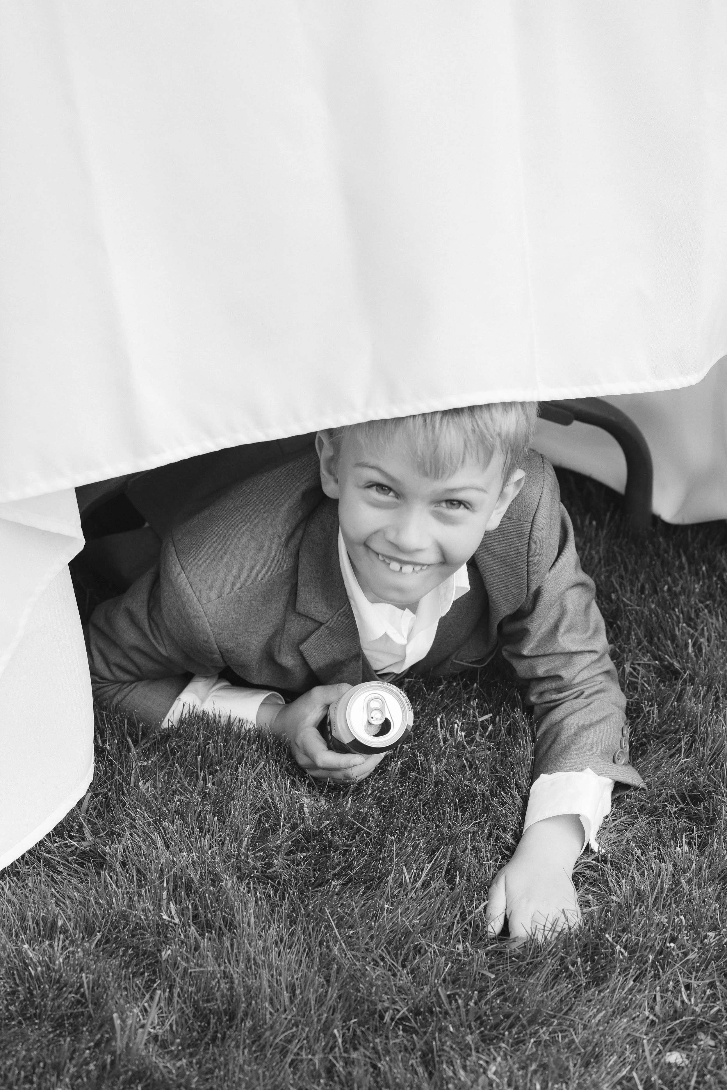 A young boy in a suit crawling on the grass under a table, holding a soda can, and smiling at the camera.
