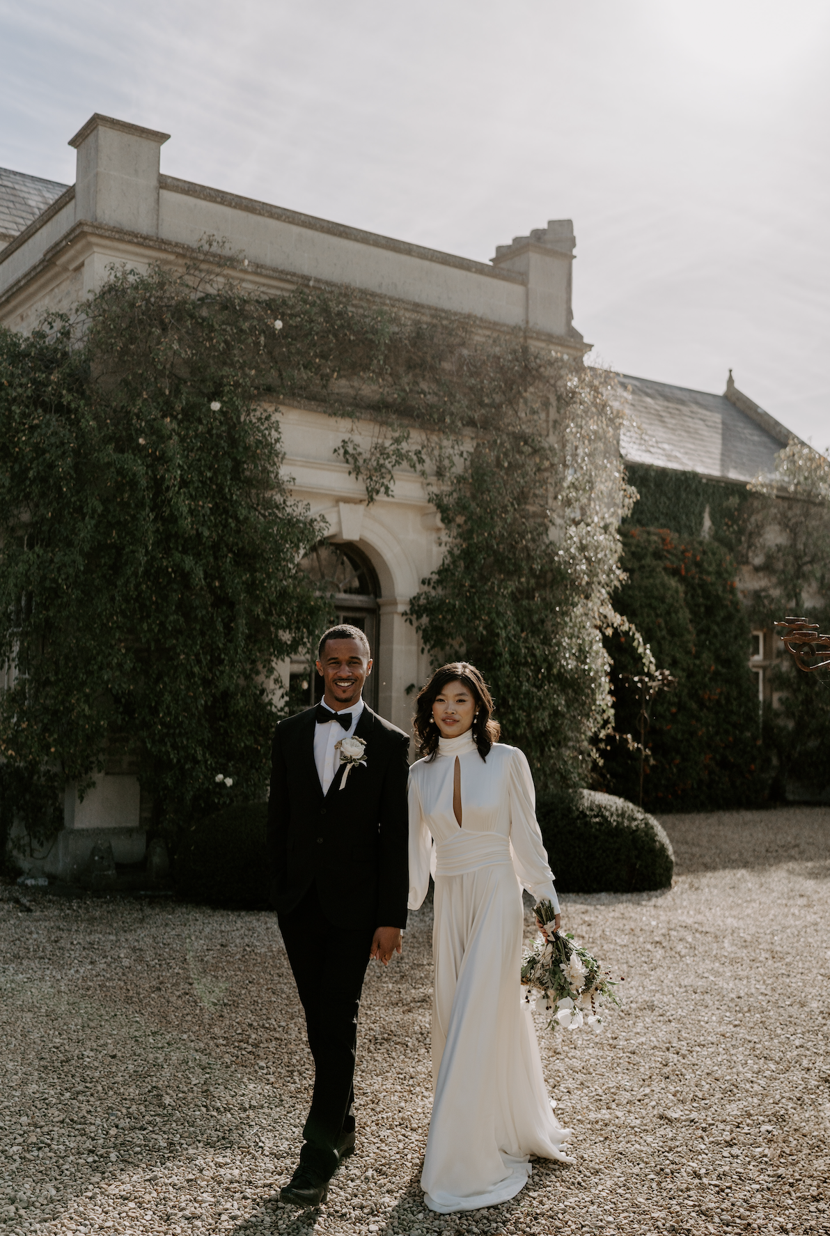 A newlywed couple walks outside a historic building, holding hands, with the bride in a white gown and the groom in a black tuxedo.