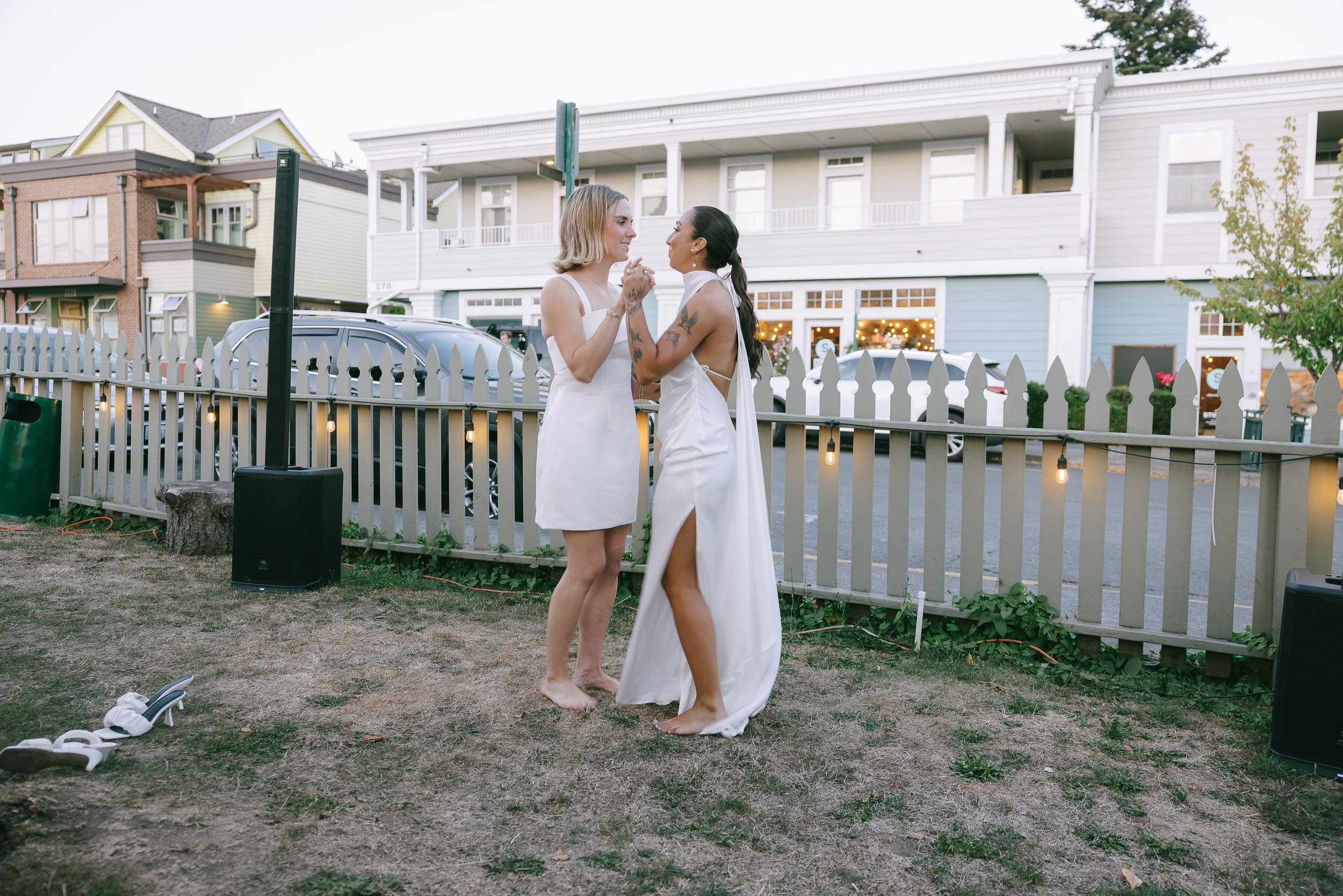 Two women in white dresses, holding hands and gazing at each other, standing on a grassy area with a white picket fence and parked cars behind them, during sunset or early evening at an outdoor event.