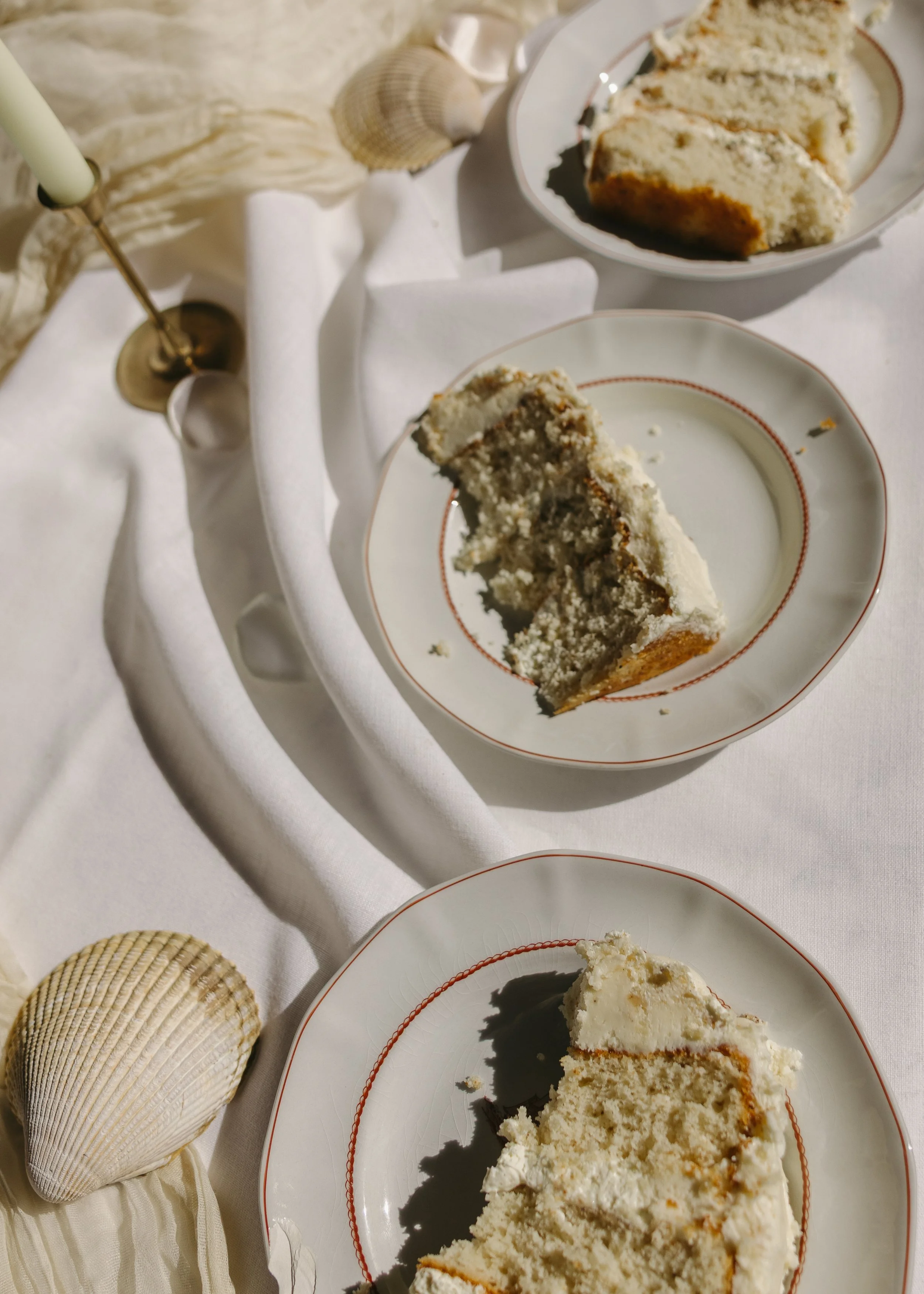 Slices of vanilla cake with white frosting on white plates, set on a table with white and beige decorations, shells, and a candle.