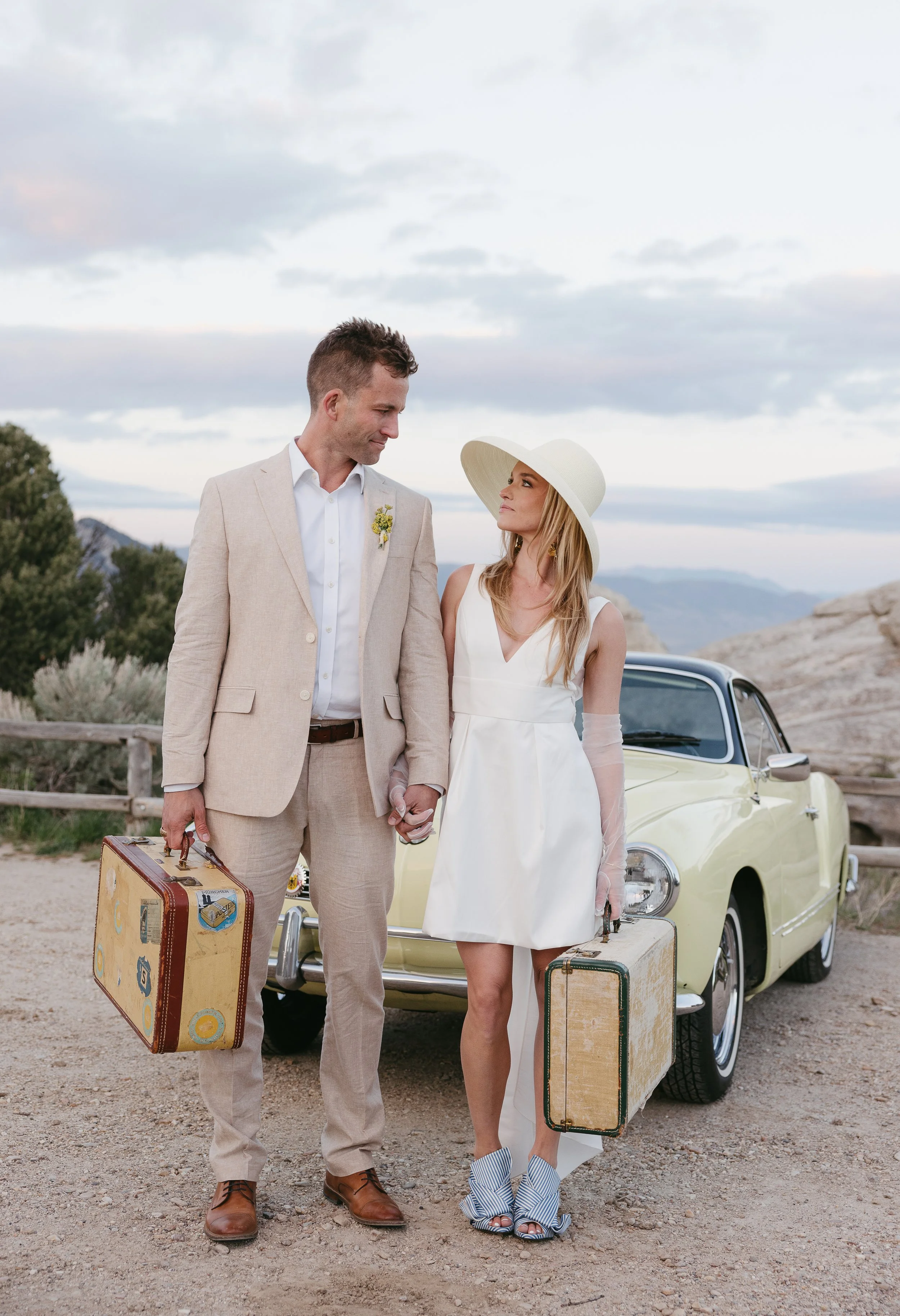 A couple dressed in vintage wedding attire standing outdoors next to a yellow classic car, holding hands, holding suitcases, with mountains and cloudy sky in the background.