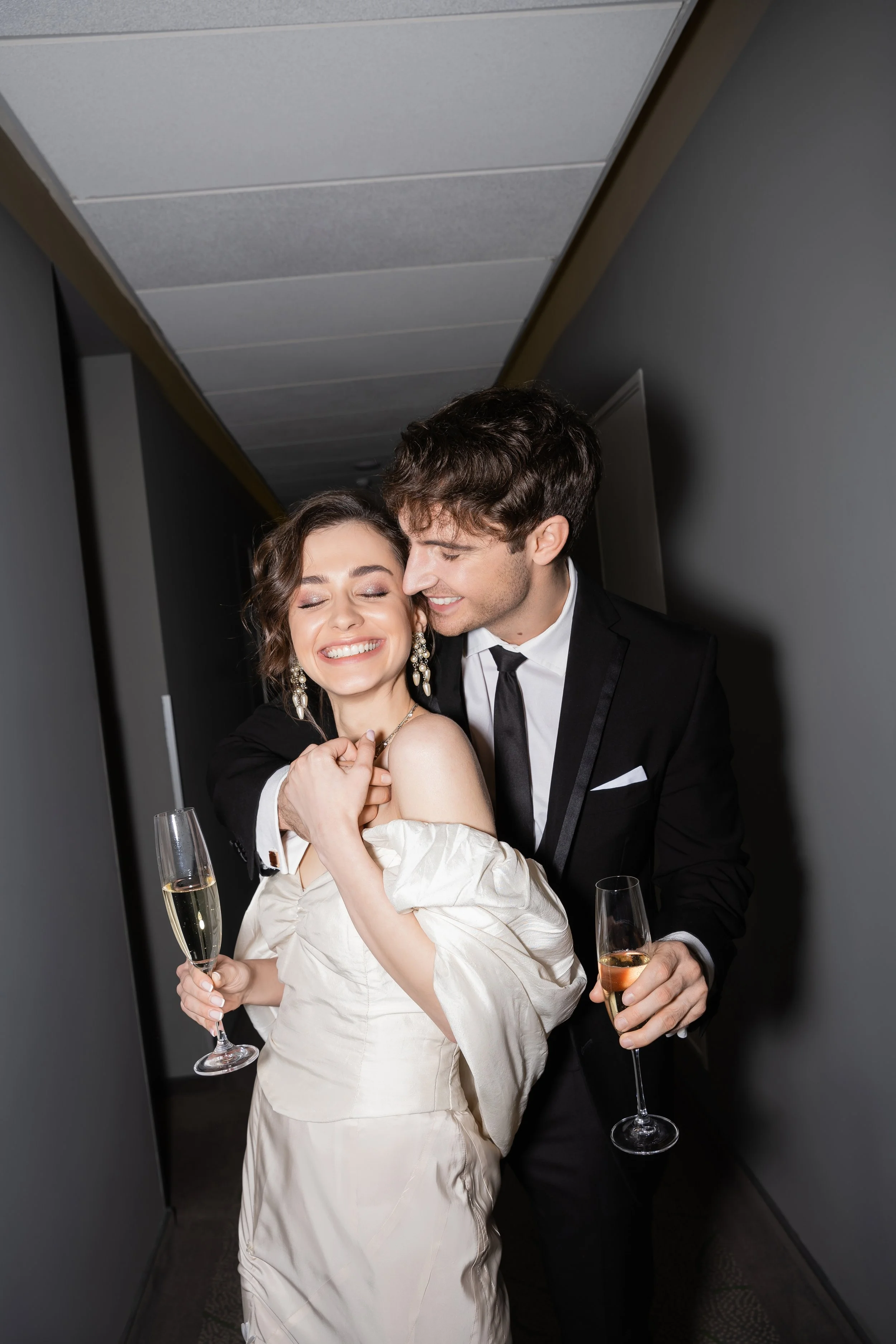 A young couple dressed in formal attire celebrating in a hall with champagne glasses, smiling joyfully.