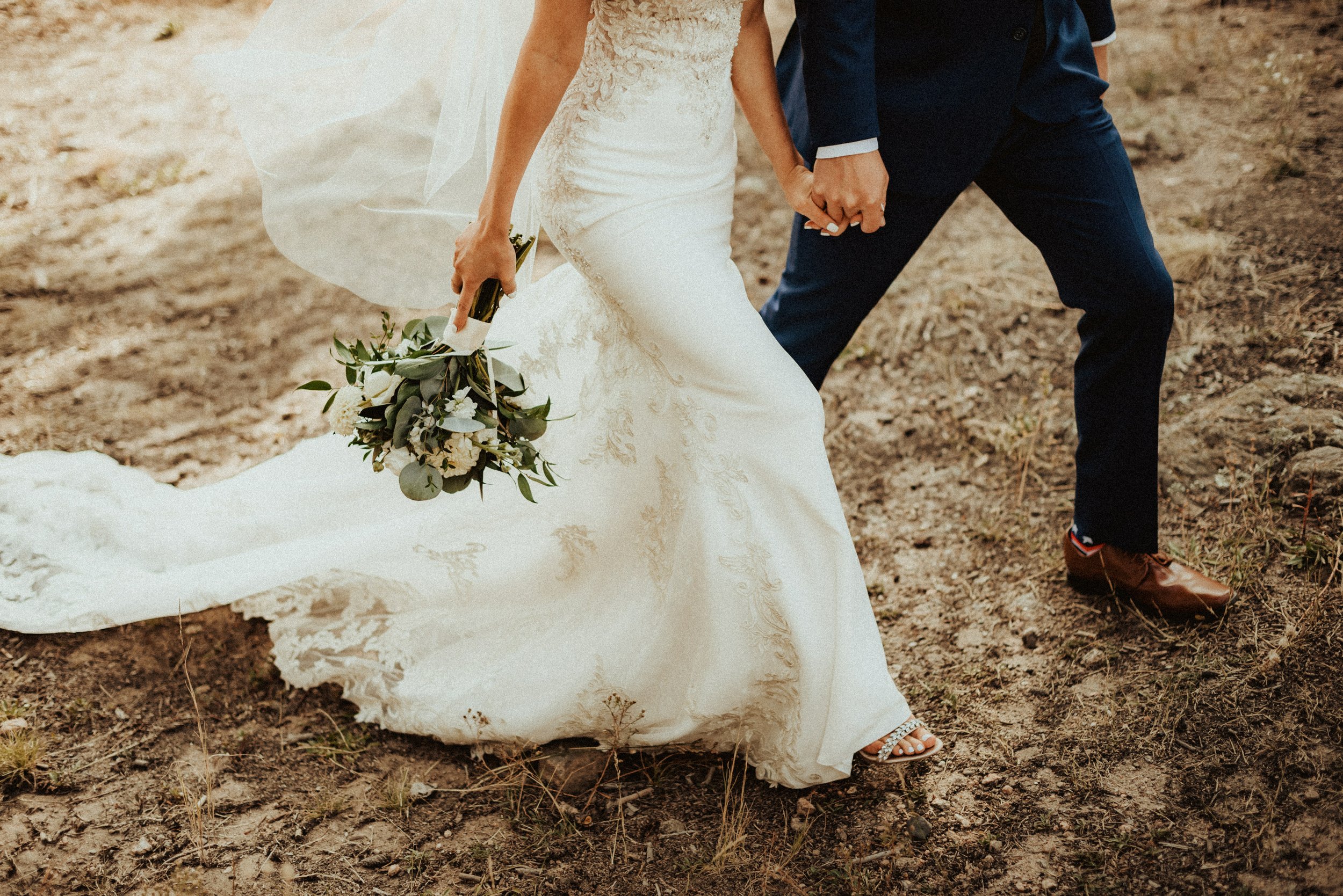 Close-up of a bride and groom holding hands walking on a dirt path during their wedding, with the bride holding a bouquet of flowers.