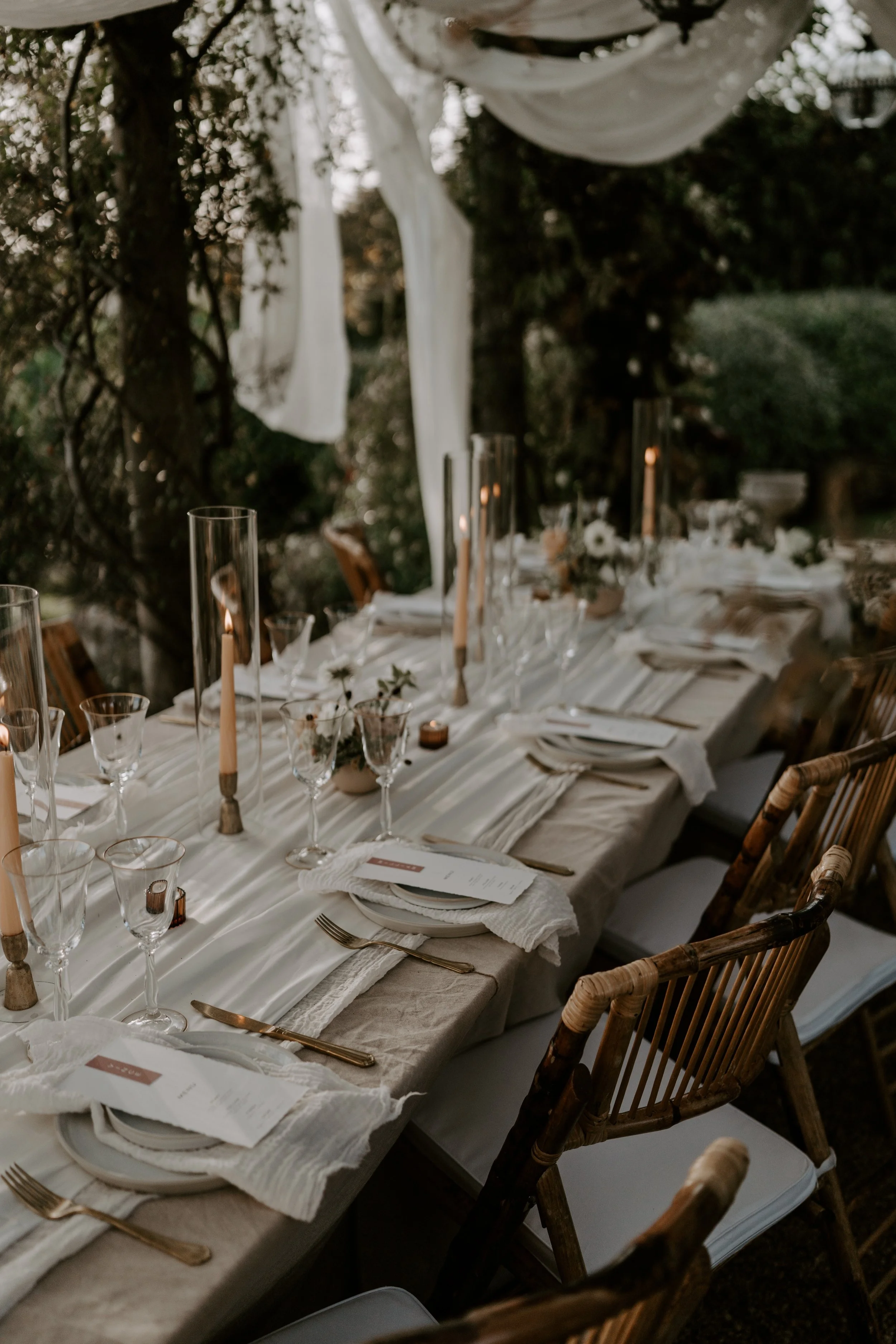 Elegant outdoor dining table set with white tablecloth, gold flatware, glassware, candles, and floral centerpieces, surrounded by wooden chairs with white cushions, overlooking a garden.