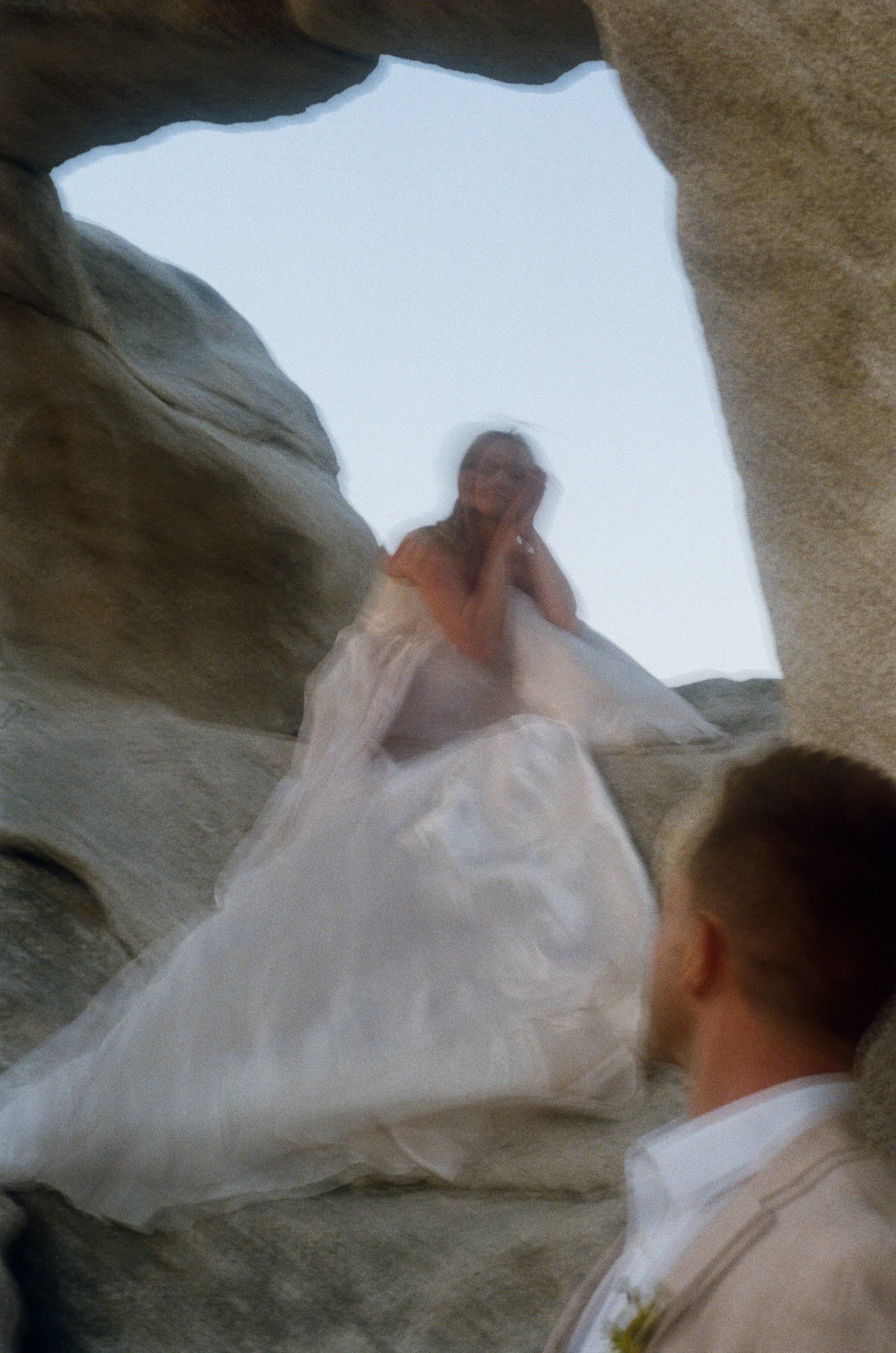 A woman in a white dress sitting between rocks with a man in a beige blazer looking at her.