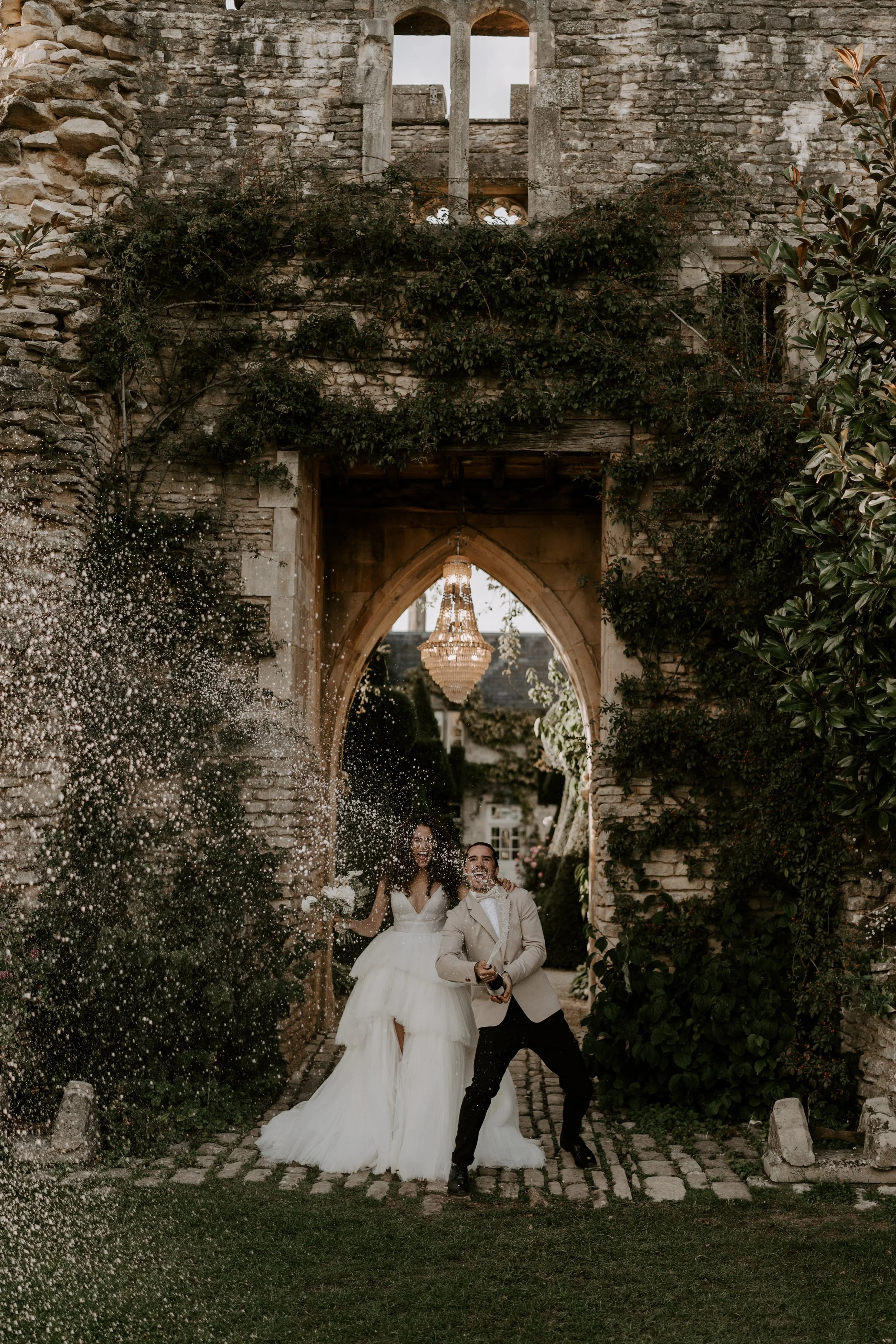 A newlywed couple celebrating behind a wedding arch with a chandelier, surrounded by greenery and a stone wall, as they throw rice.