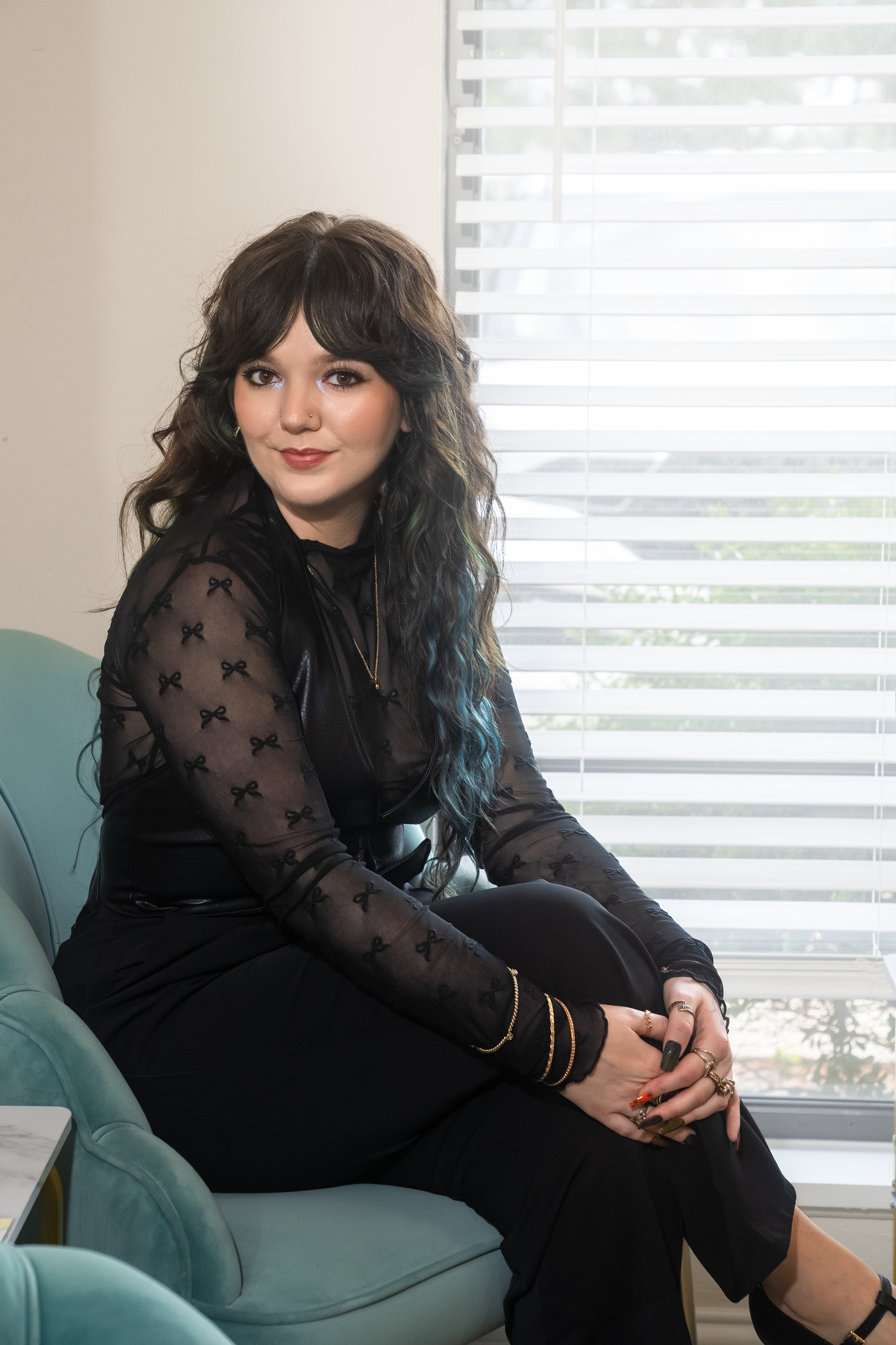 A woman with brown and teal hair sitting in a chair, smiling.
