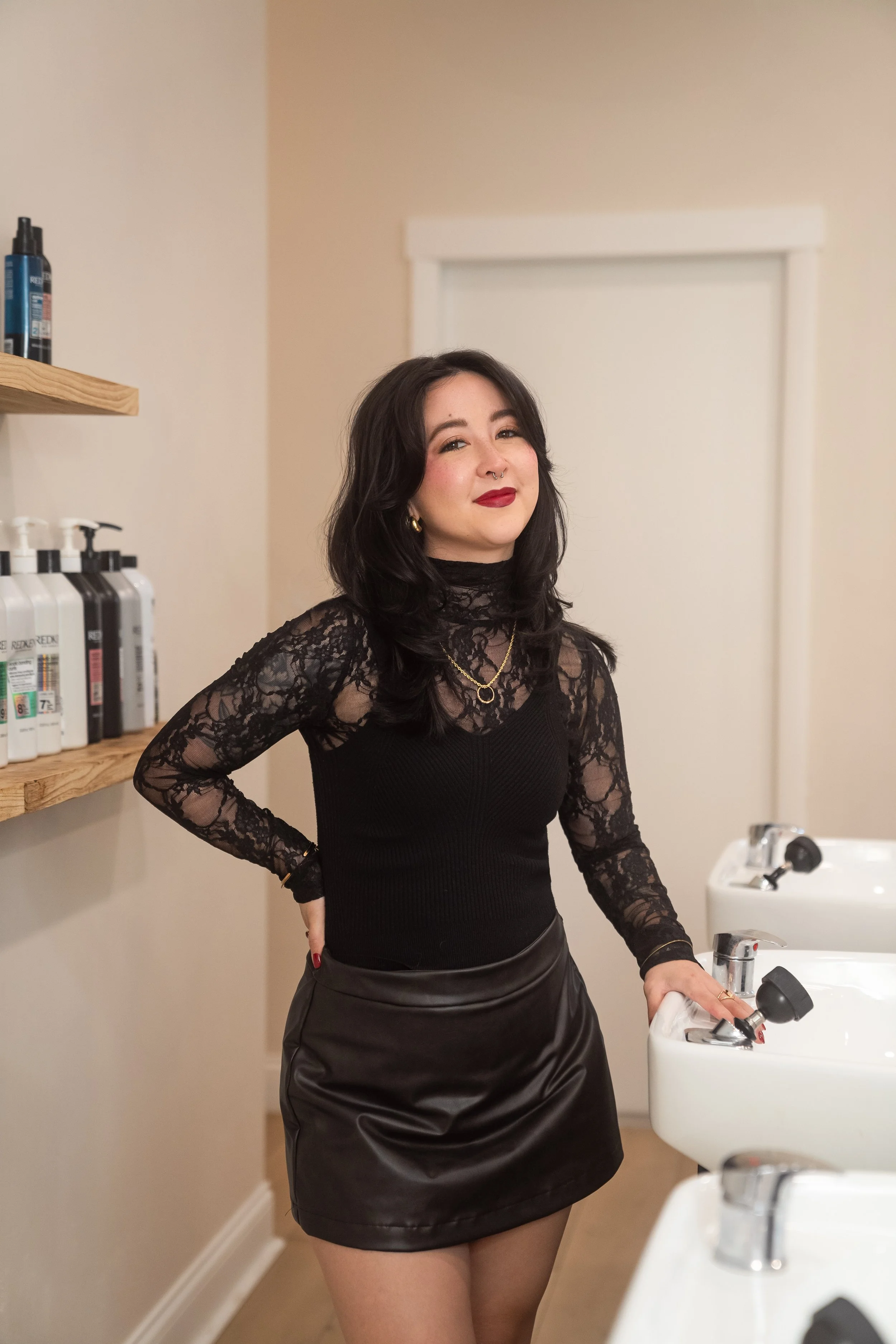 Image is of a woman with chest length dark drown hair standing next to a shampoo bowl, smiling.