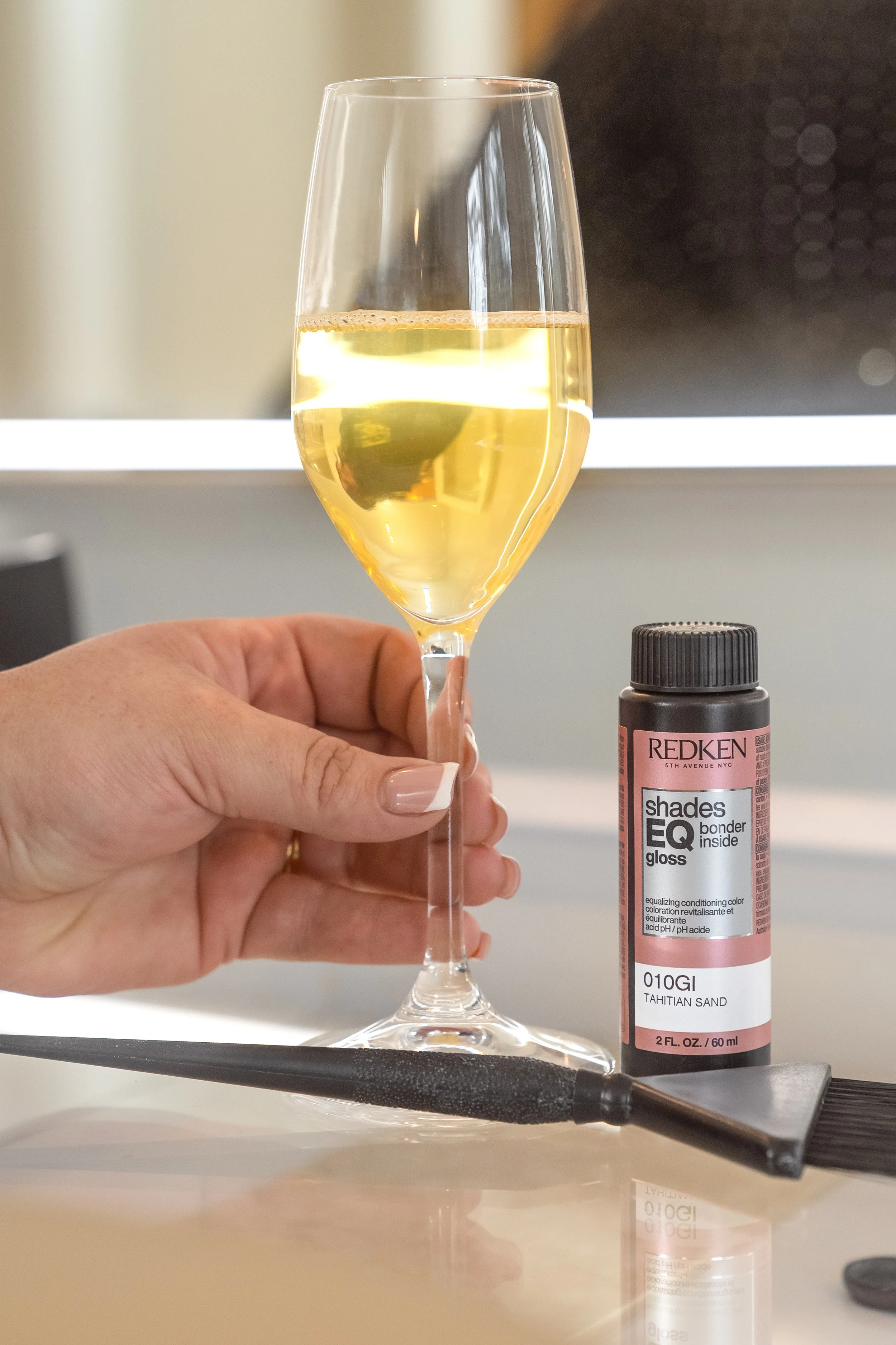Image is of a hand holding a glass of champagne with a bottle of Redken Toner next to it.