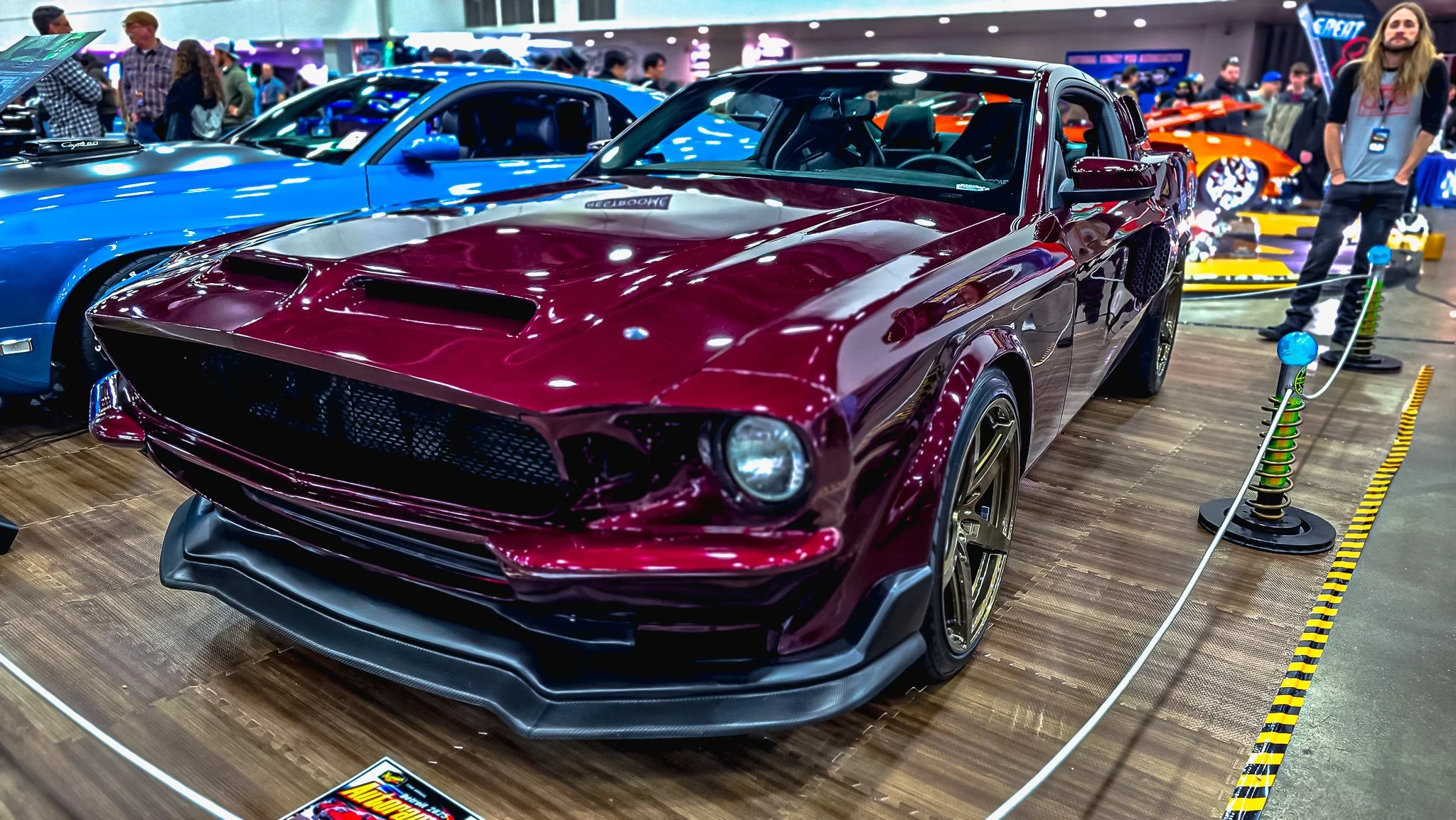 A classic race car with a maroon body on display at an auto show, surrounded by a crowd of people and other vehicles.