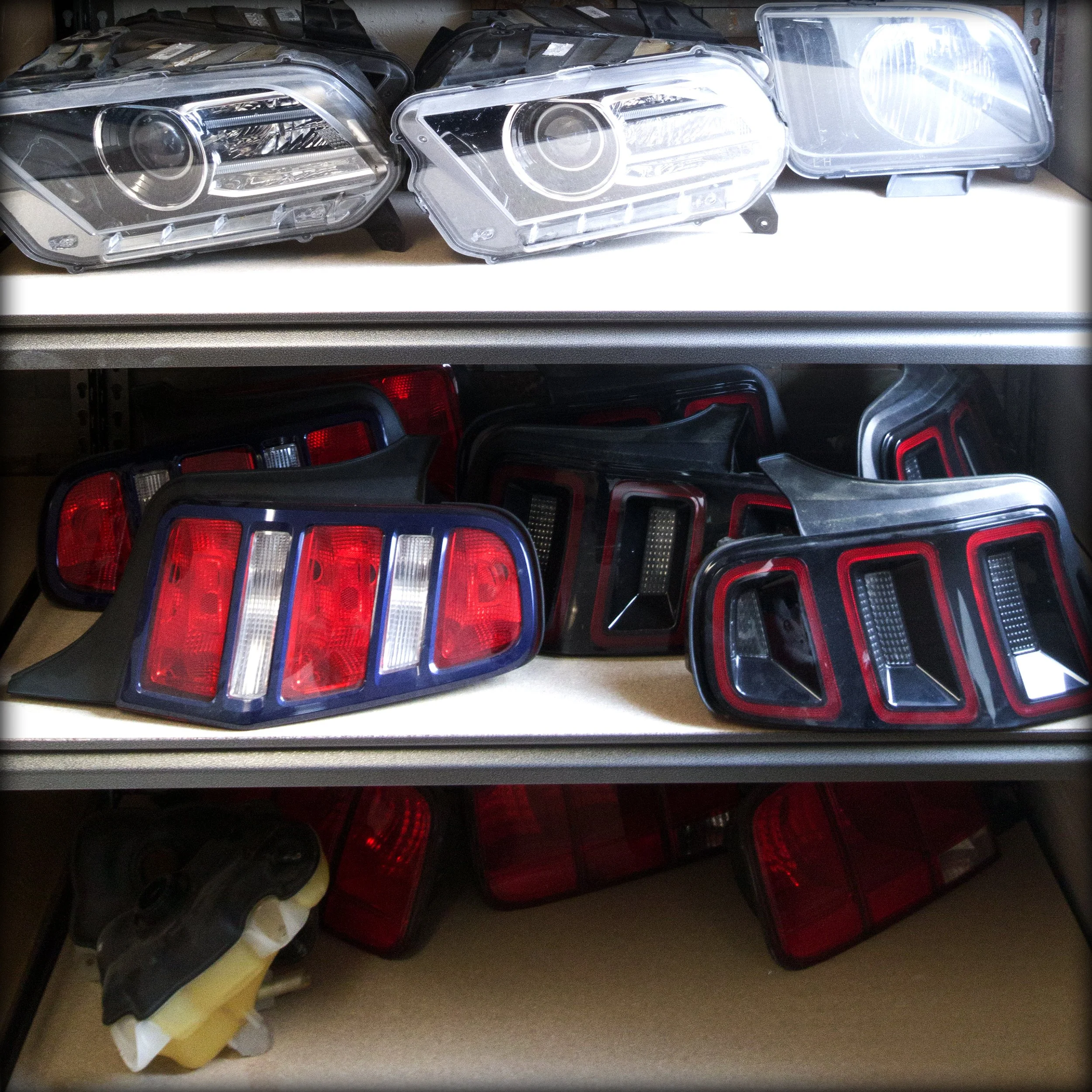 Three shelves filled with automotive tail lights and headlight components, including red and clear lenses and a fuel reservoir on the bottom shelf.