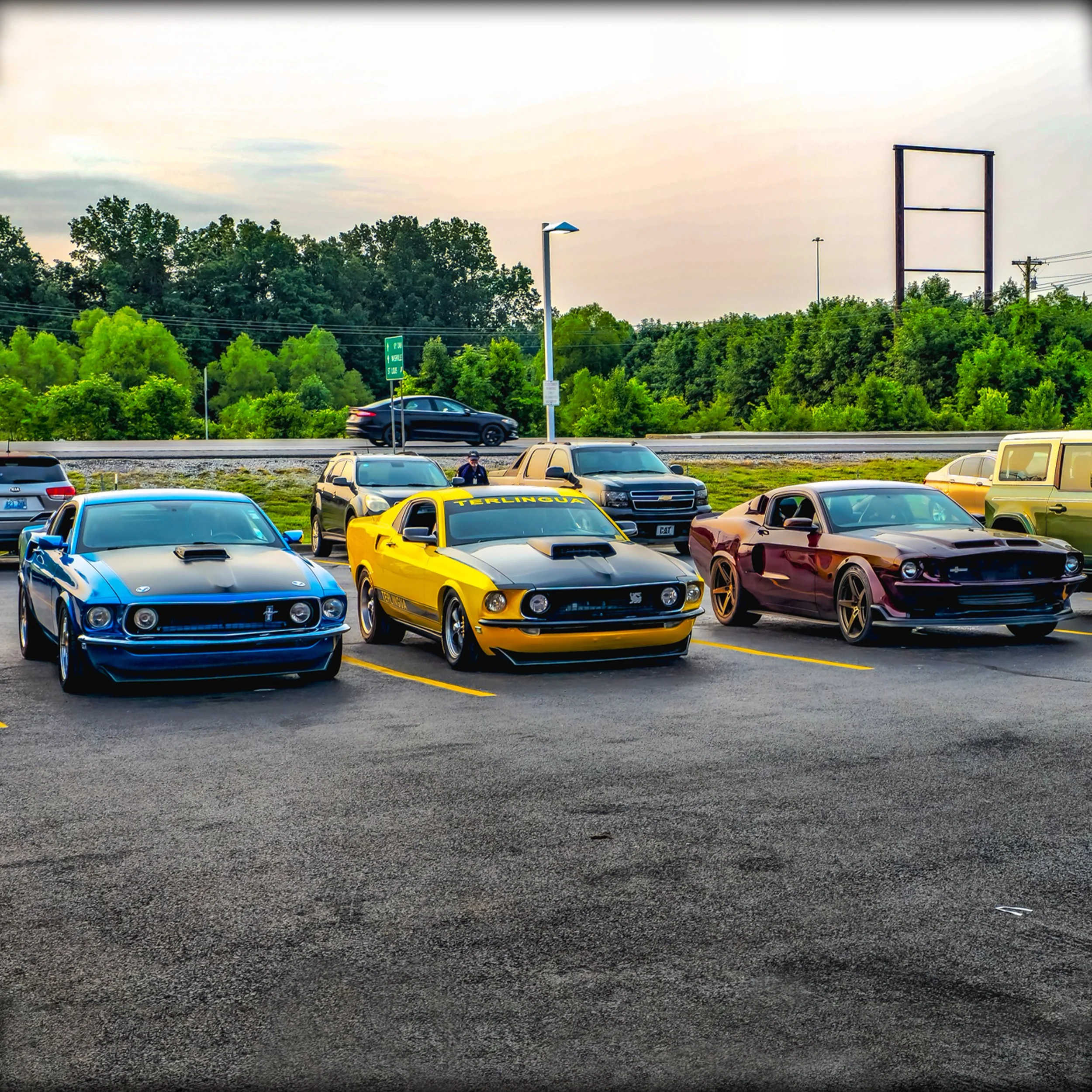 A parking lot with four classic Ford Mustangs parked side by side, with a few modern vehicles in the background, all under a partly cloudy sky.