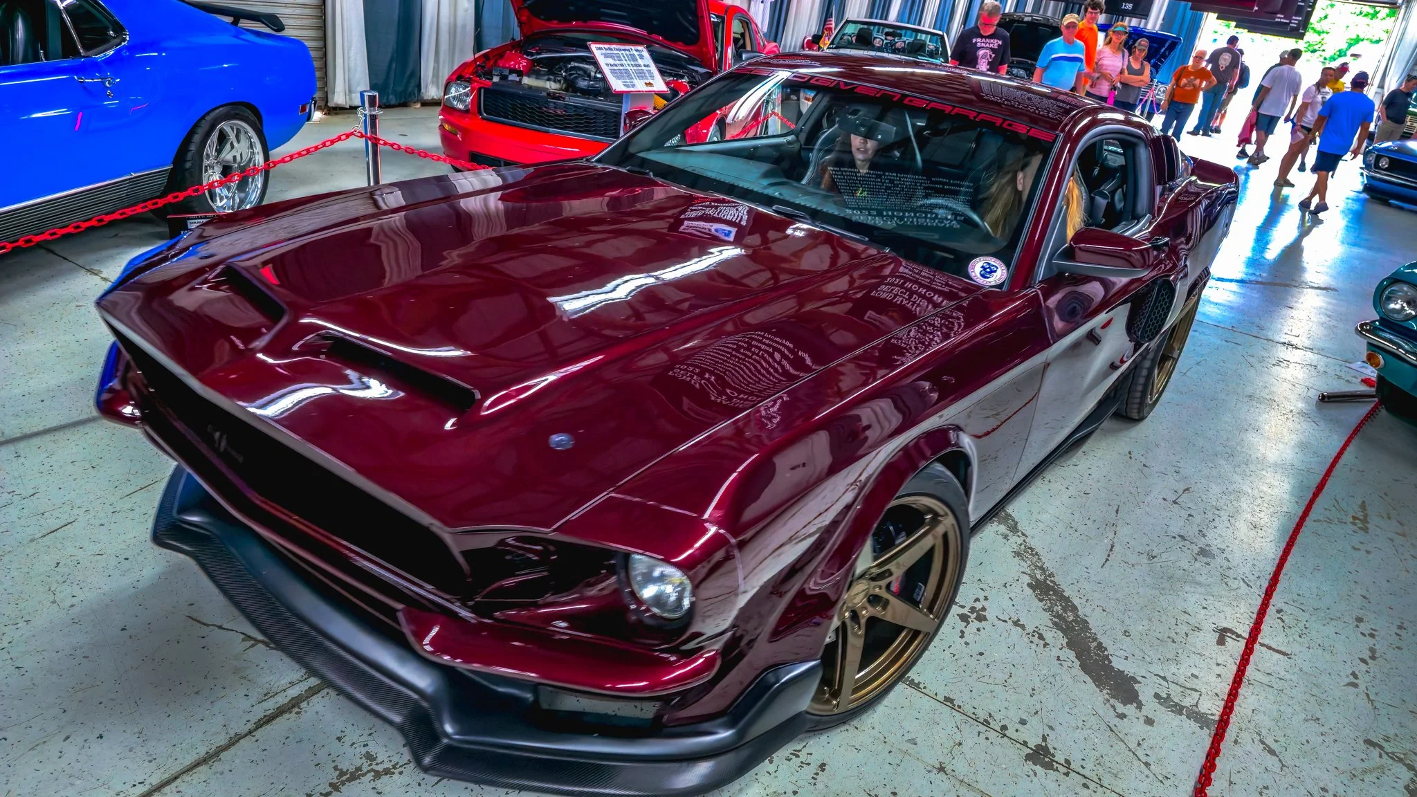 A maroon sports car with gold wheels on display at an indoor car show, surrounded by other vehicles and visitors.