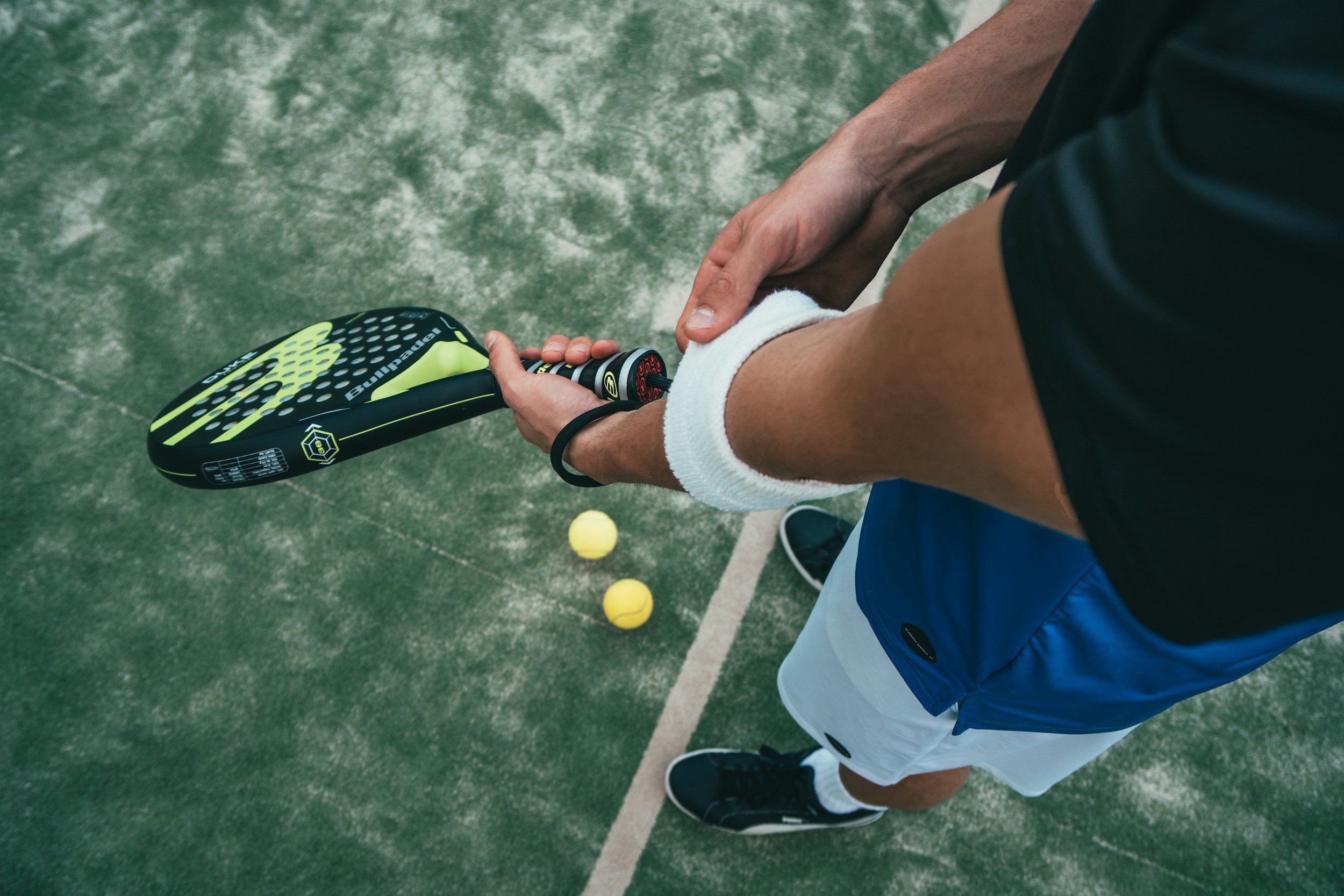 A person holding a padel racket with two yellow padel balls on a green tennis court.