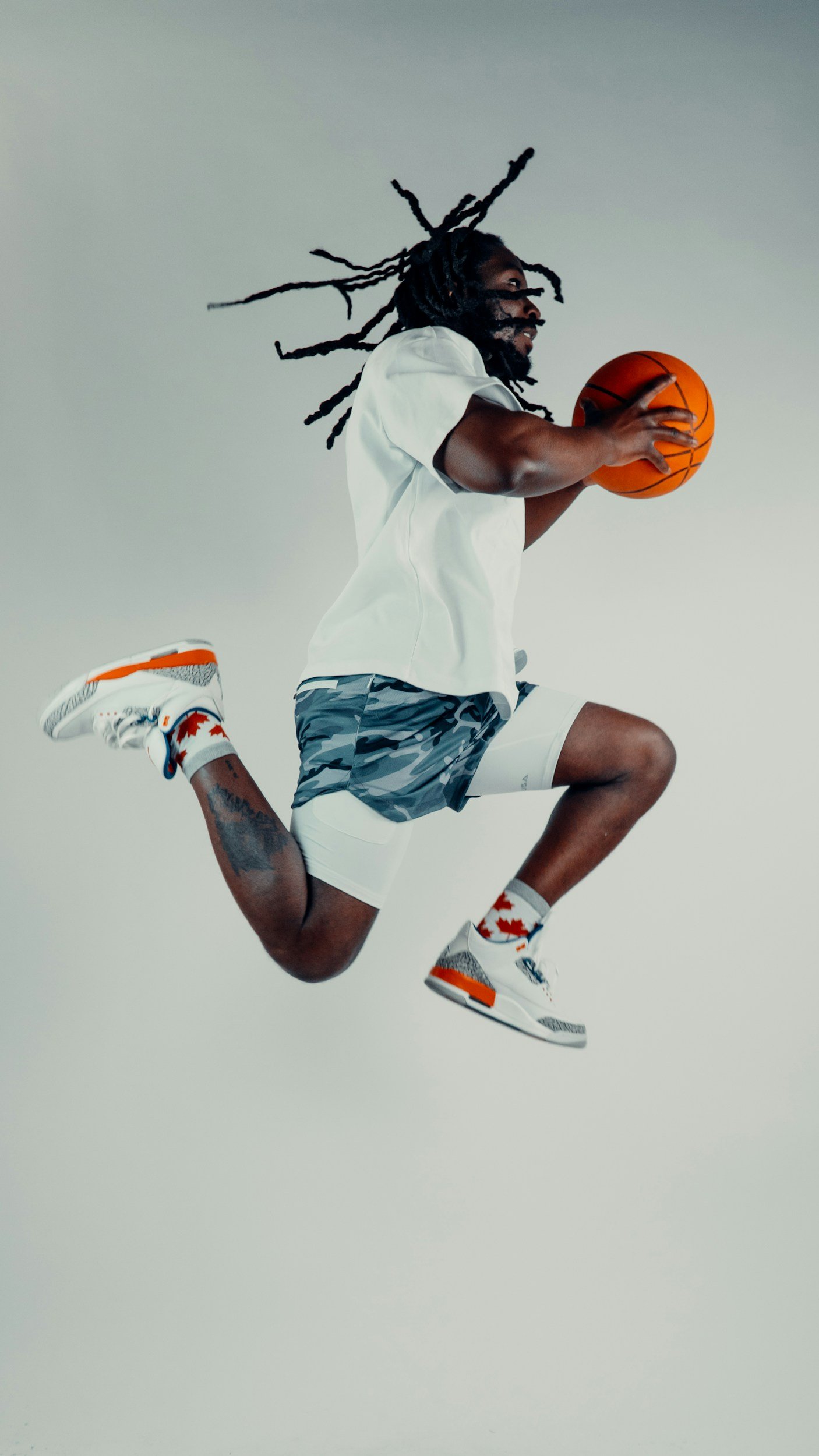 A man with long dreadlocks jumping in the air while holding an orange basketball, wearing a white t-shirt, camouflage shorts, and white sneakers with red and black accents.