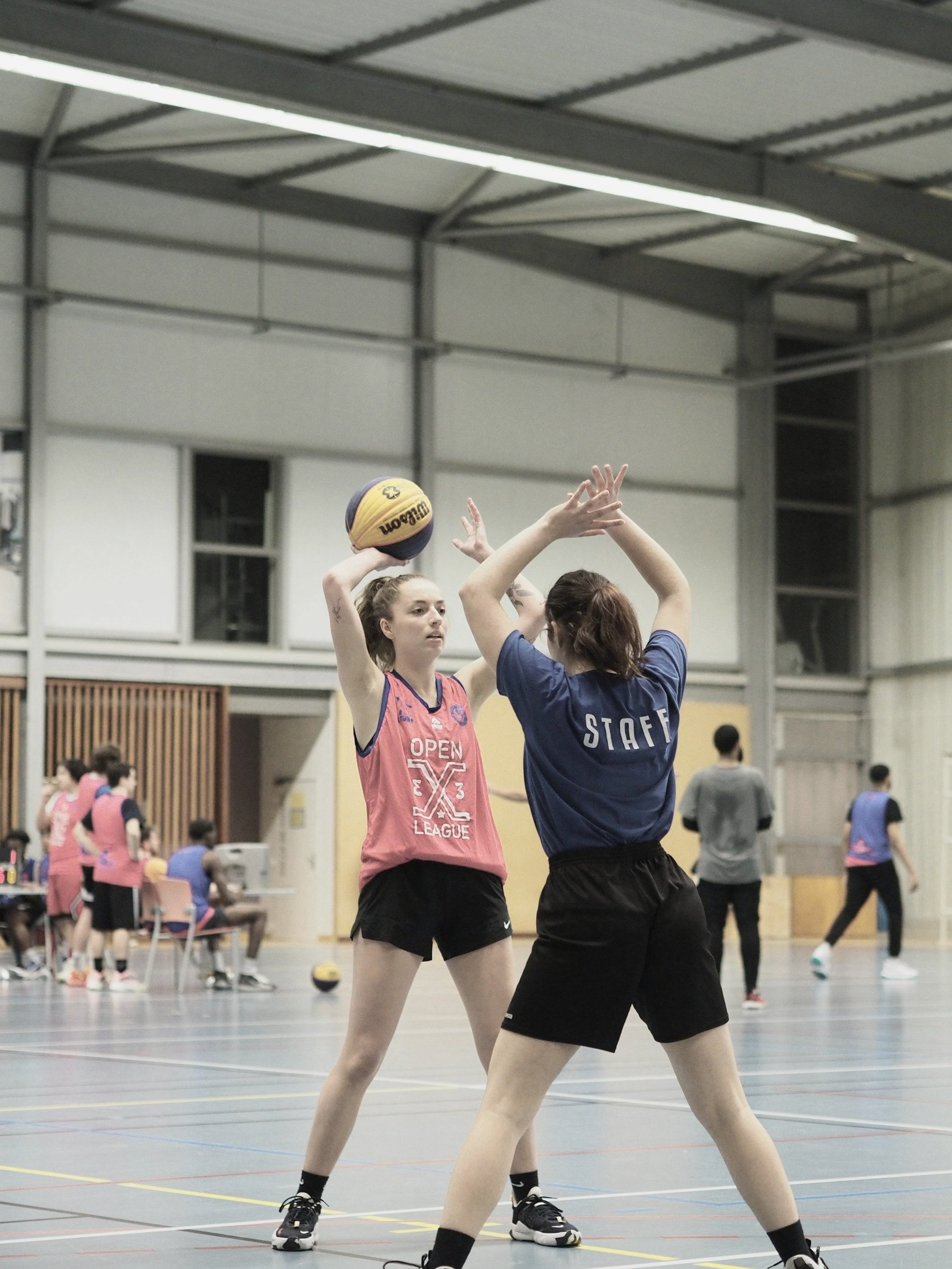 A female volleyball player in a pink jersey preparing to hit a volleyball while a staff member in a blue shirt stands in front of her on an indoor court.