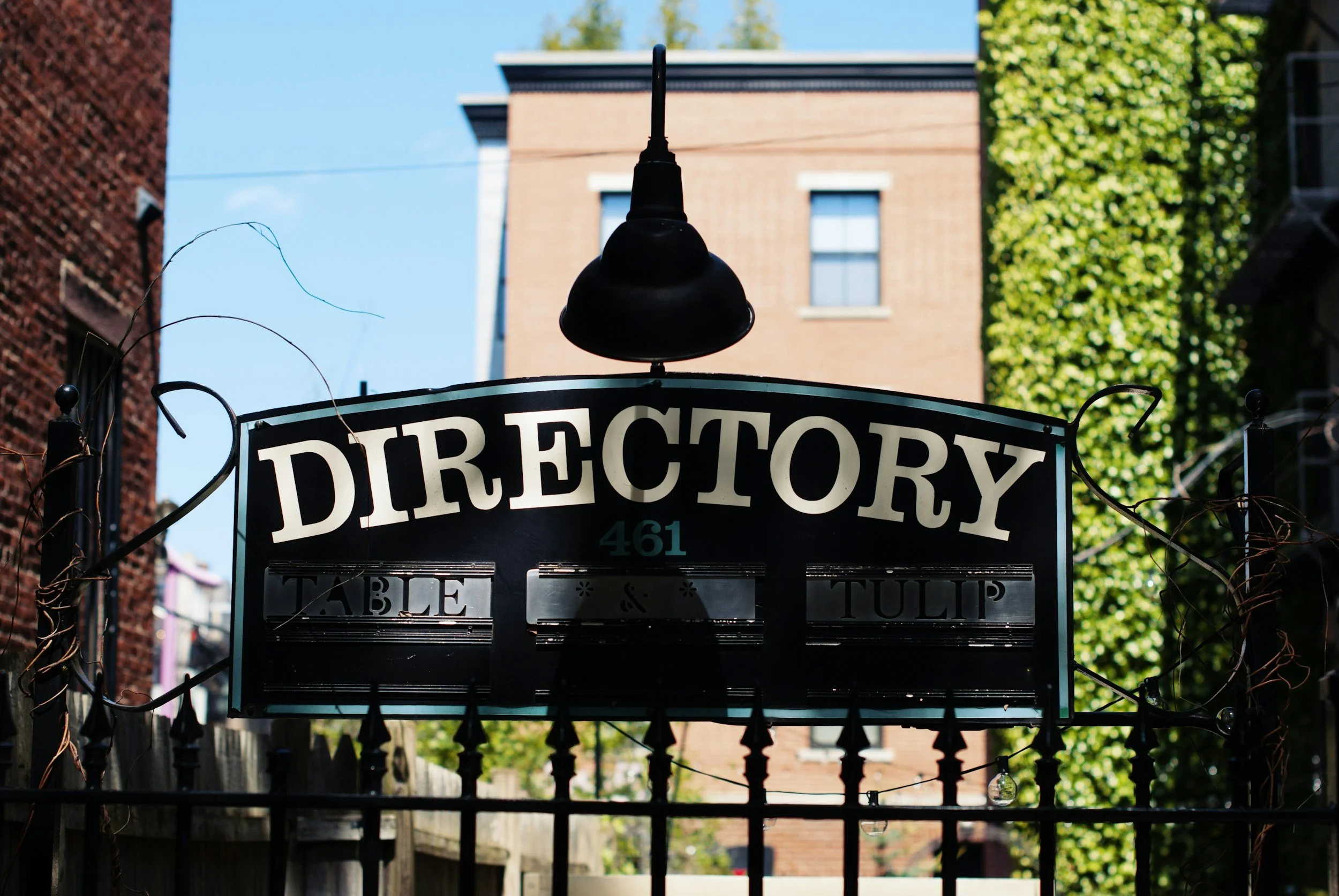 A black metal gate with a sign that reads 'Drectory' and a black lamp post on top of the gate, with a brick building and green trees in the background.
