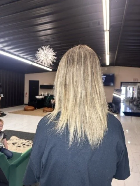 Back of a woman with shoulder-length blonde hair, standing in an indoor space with black ceiling and bright lights, with Christmas decorations hanging from the ceiling, and a green sofa to her left.