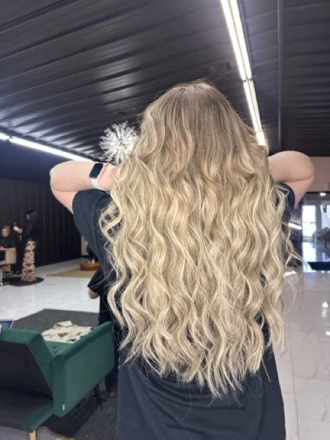 Woman with long, wavy blonde hair standing indoors with her back to the camera, arms resting on her head.