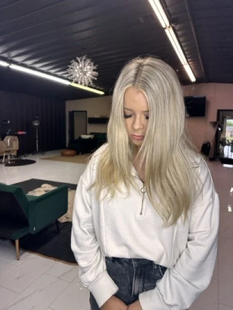 Young woman with long blonde hair wearing a white sweatshirt and dark pants, standing in a modern salon or basement with black ceiling and neon lights.