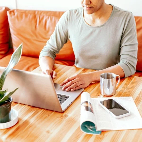 Person working on a laptop at a wooden table, with a smartphone, a magazine, a metal mug, and a potted plant nearby.