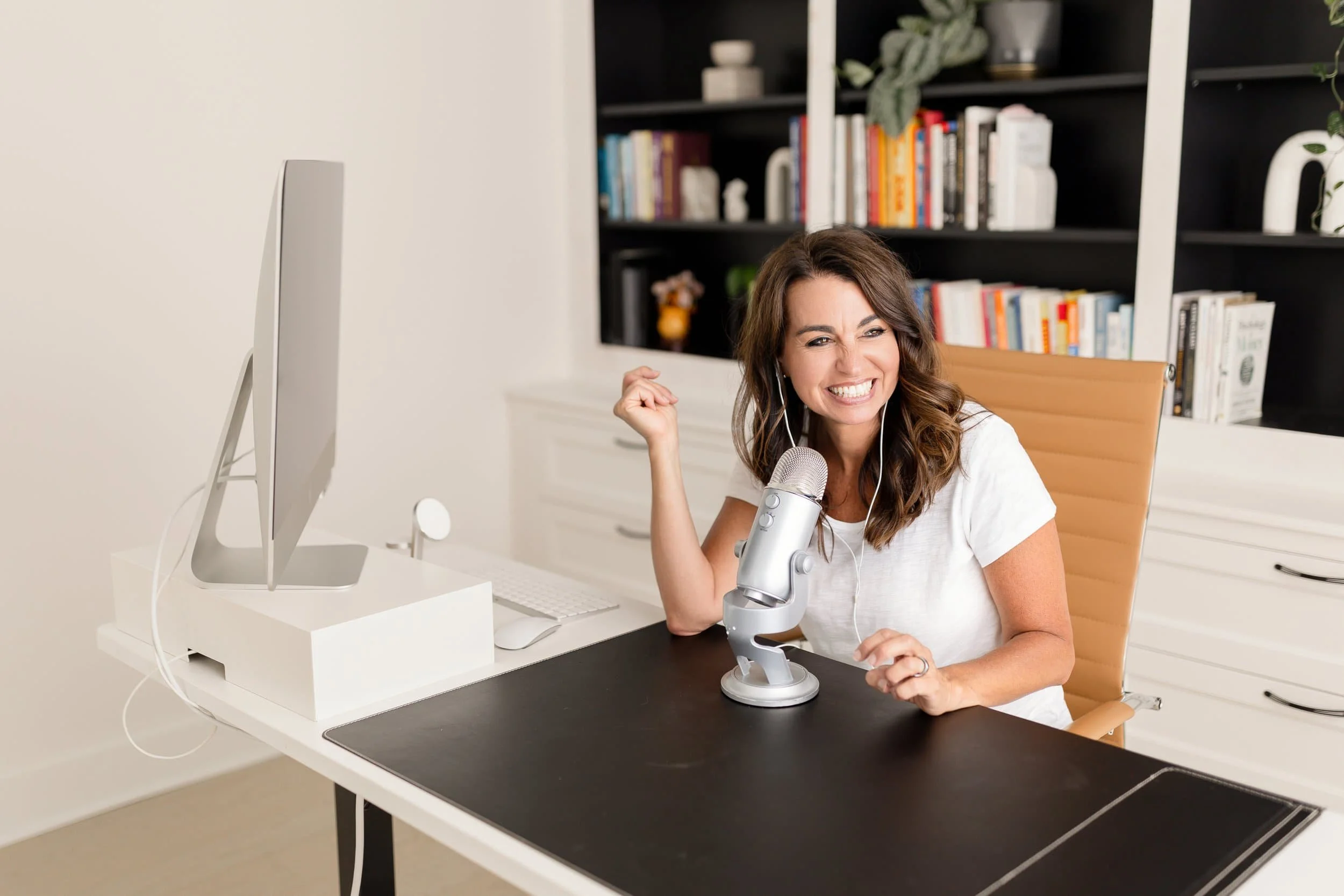 Smiling woman with earphones sitting at desk with microphone in front, computer monitor to her left, and bookshelf behind her.