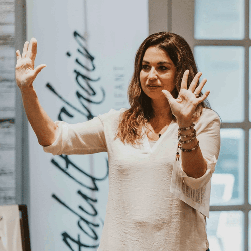 A woman with shoulder-length brown hair, wearing a white blouse, gestures with both hands raised, palms facing outward, as she speaks in front of a glass window and a sign in the background.