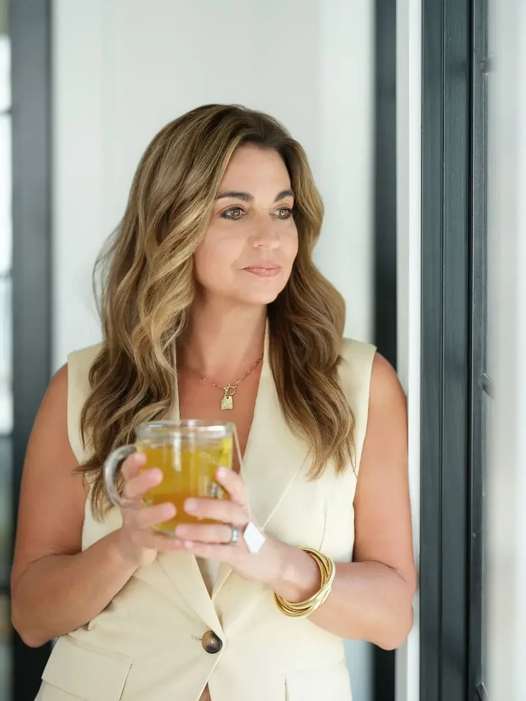 A woman with wavy brown hair holding a glass mug of tea standing near a window.