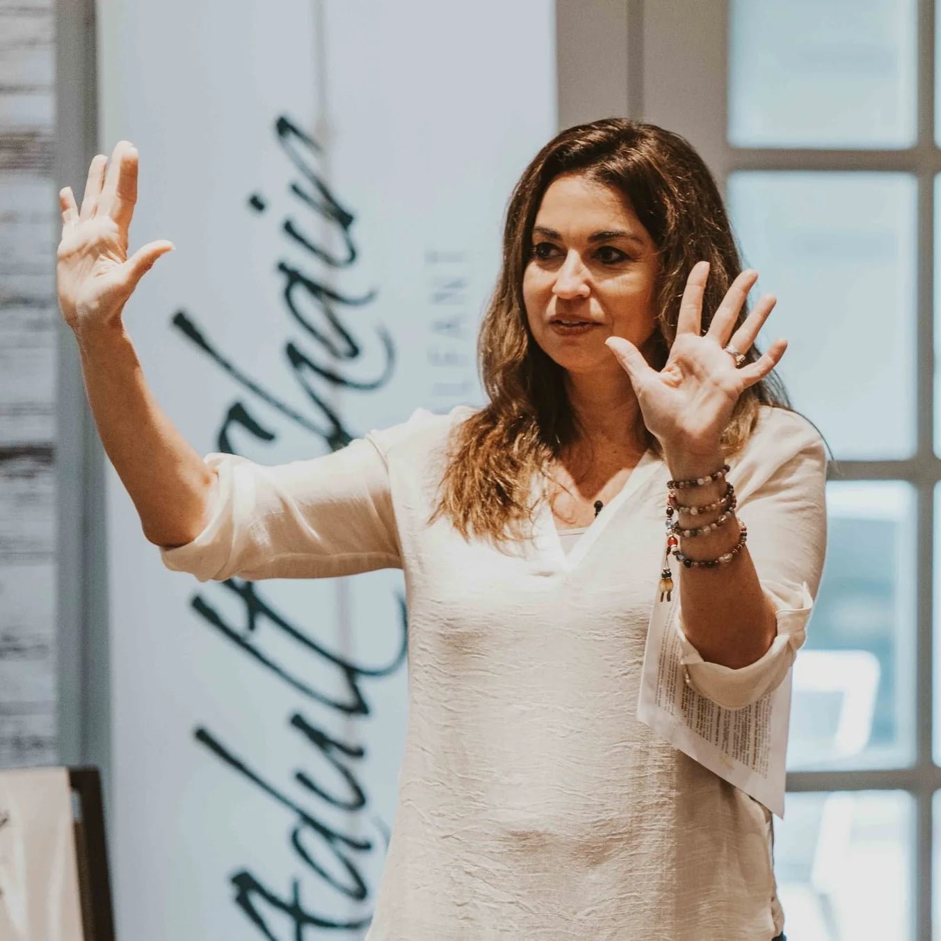 A woman in a white blouse gestures with both hands raised during a presentation in a bright room with large windows.