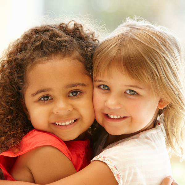 Two young girls hugging and smiling at the camera indoors, with sunlight coming through a window in the background.
