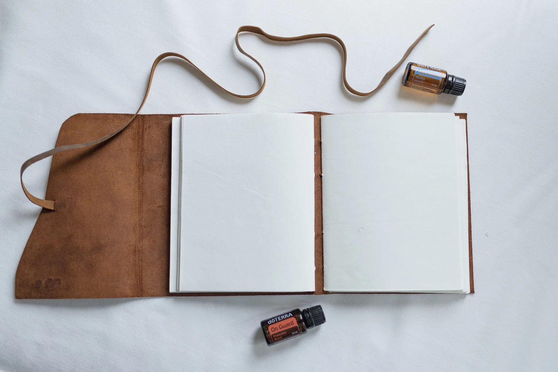 Open notebook with blank pages on a brown leather cover, two bottles of doTERRA essential oils labeled "On Guard" and "Ginger" on a white surface.