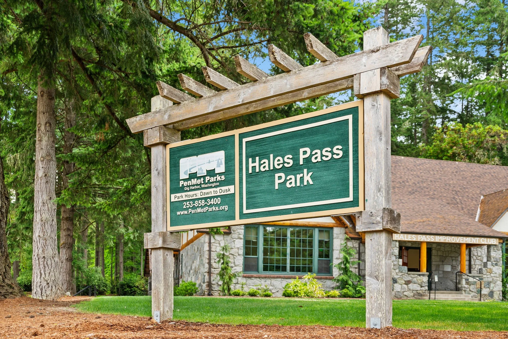 Sign at Hales Pass Park in PenMet Parks, Gig Harbor, Washington, with a stone building and tall trees in the background.