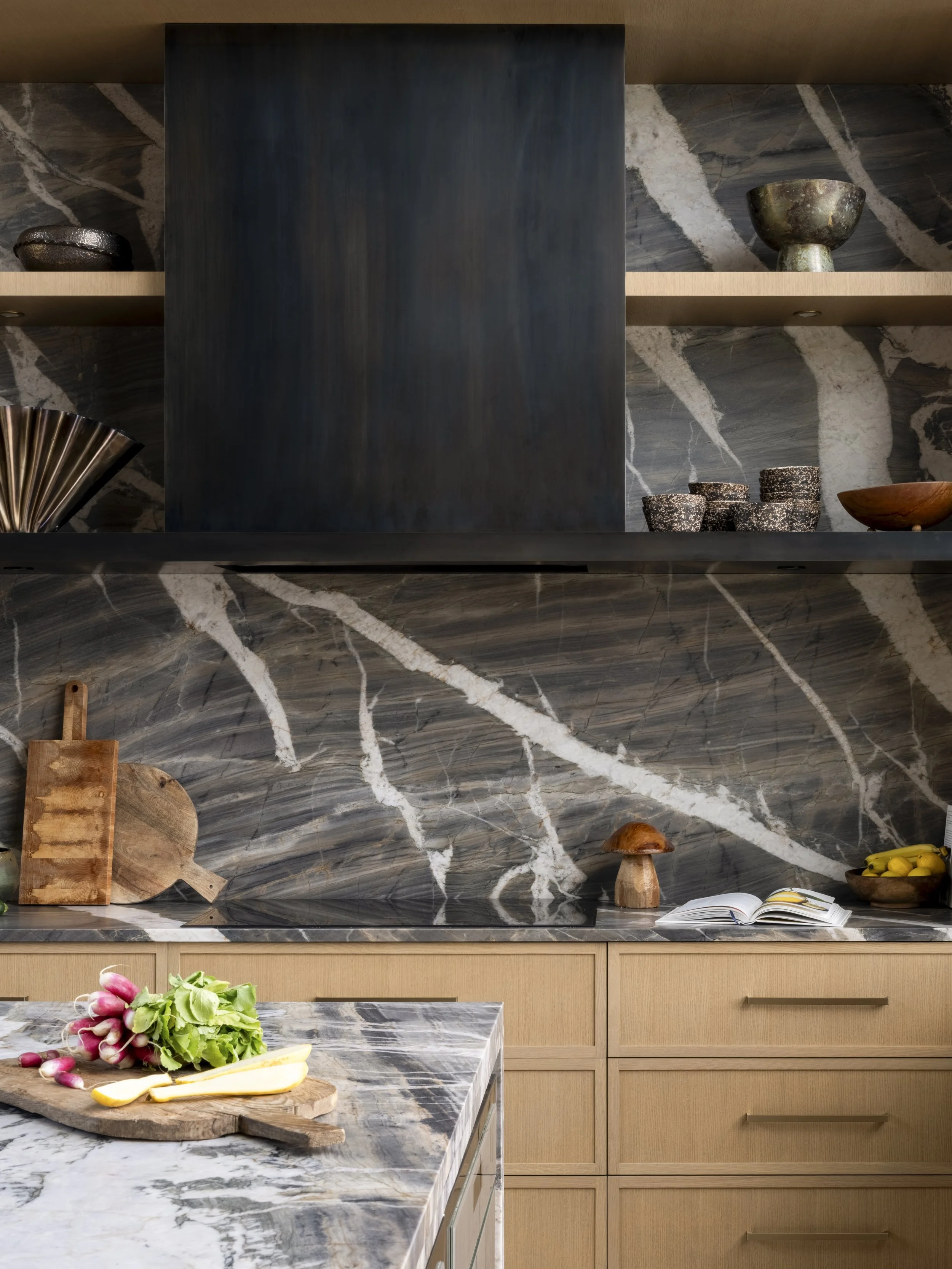 Kitchen with marble backsplash, open shelves, and wooden cabinets, featuring vegetables, mushrooms, chopping boards, and a cookbook.