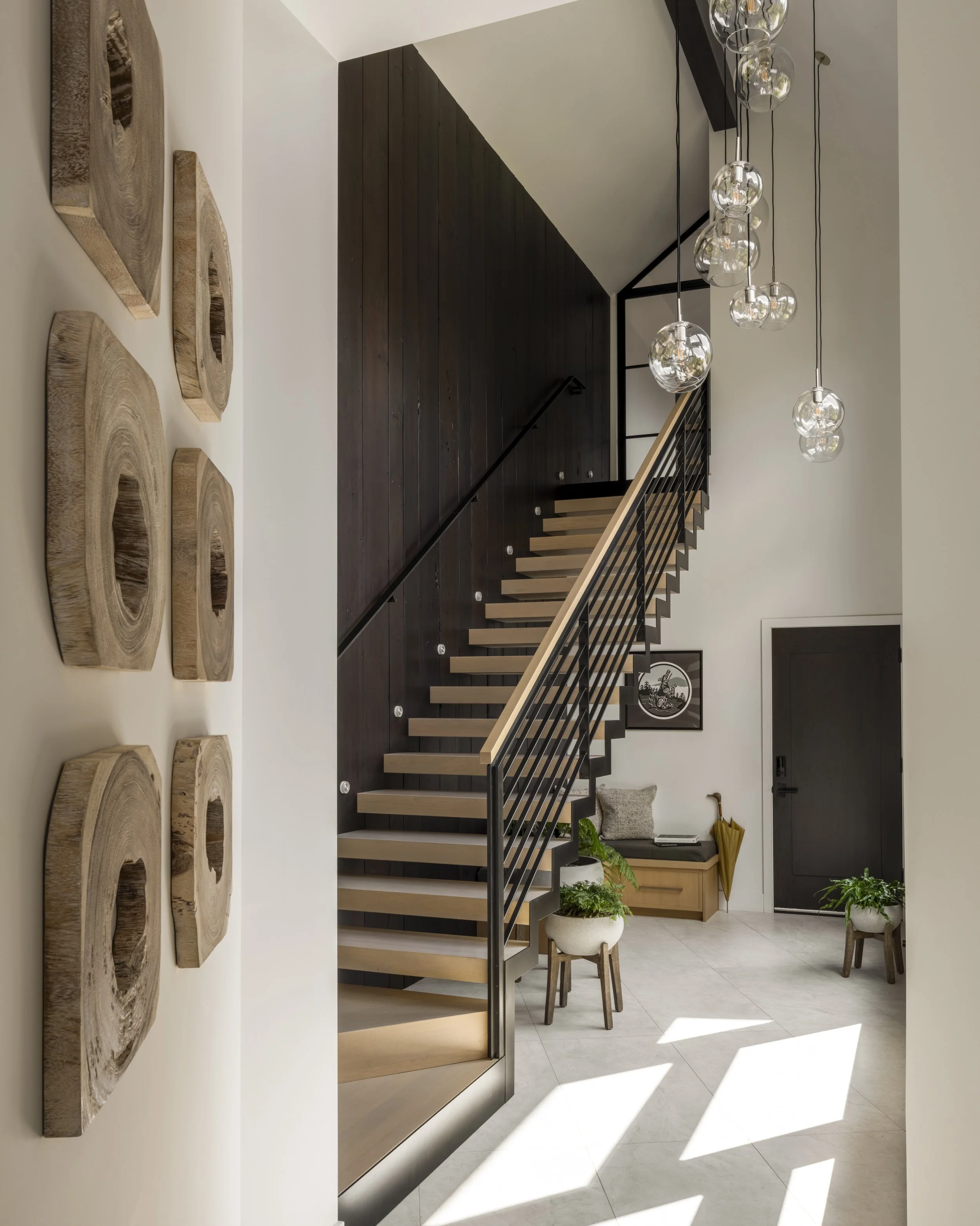 Modern staircase with wooden steps and black metal railing against dark wood-paneled wall in a bright entryway with white walls, decorative wall art, hanging glass globe light fixtures, potted plants, and a small bench.