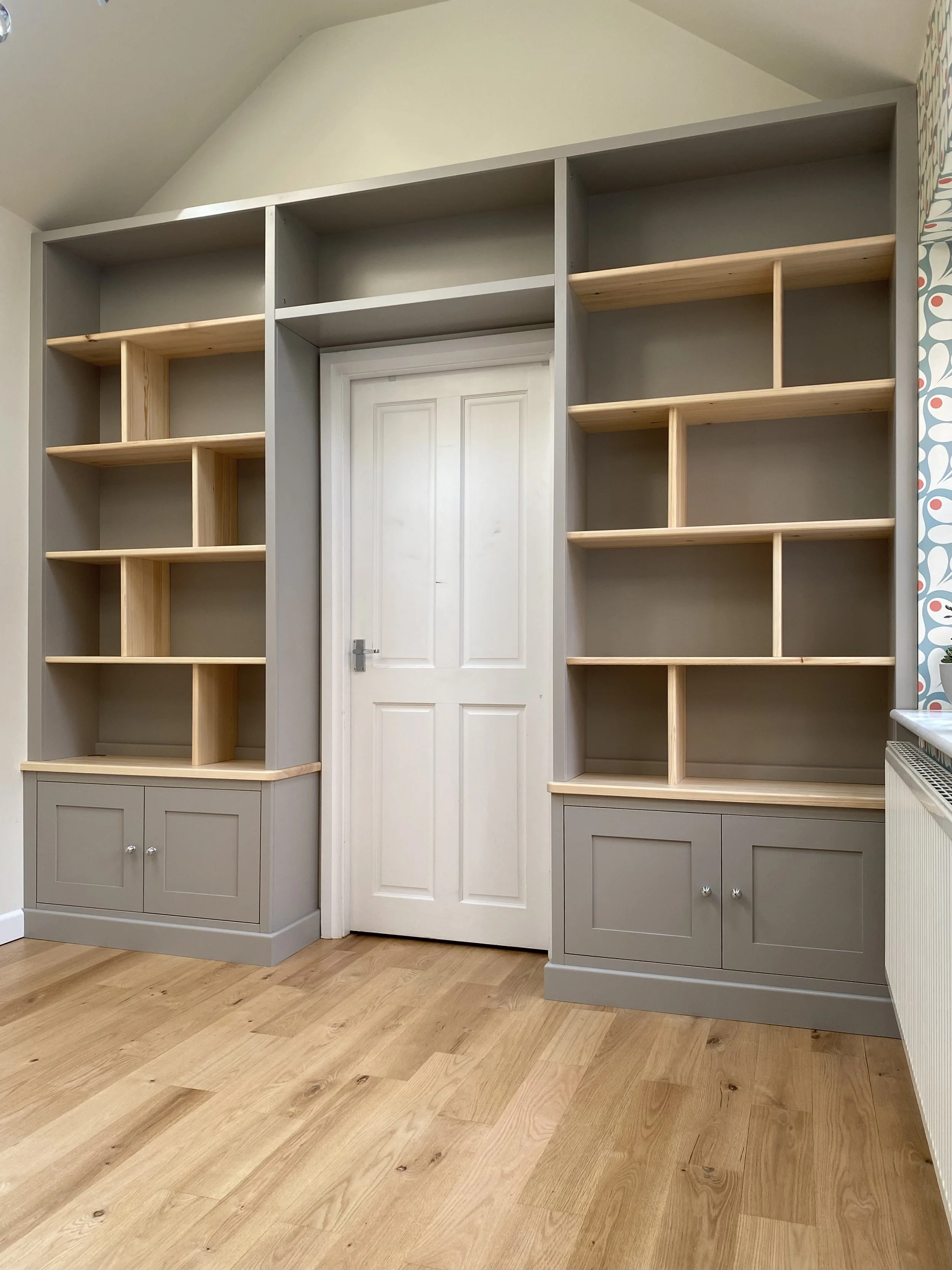 Empty built-in bookshelves with a door in the middle, painted grey with natural wood shelves, in a room with light wood flooring.
