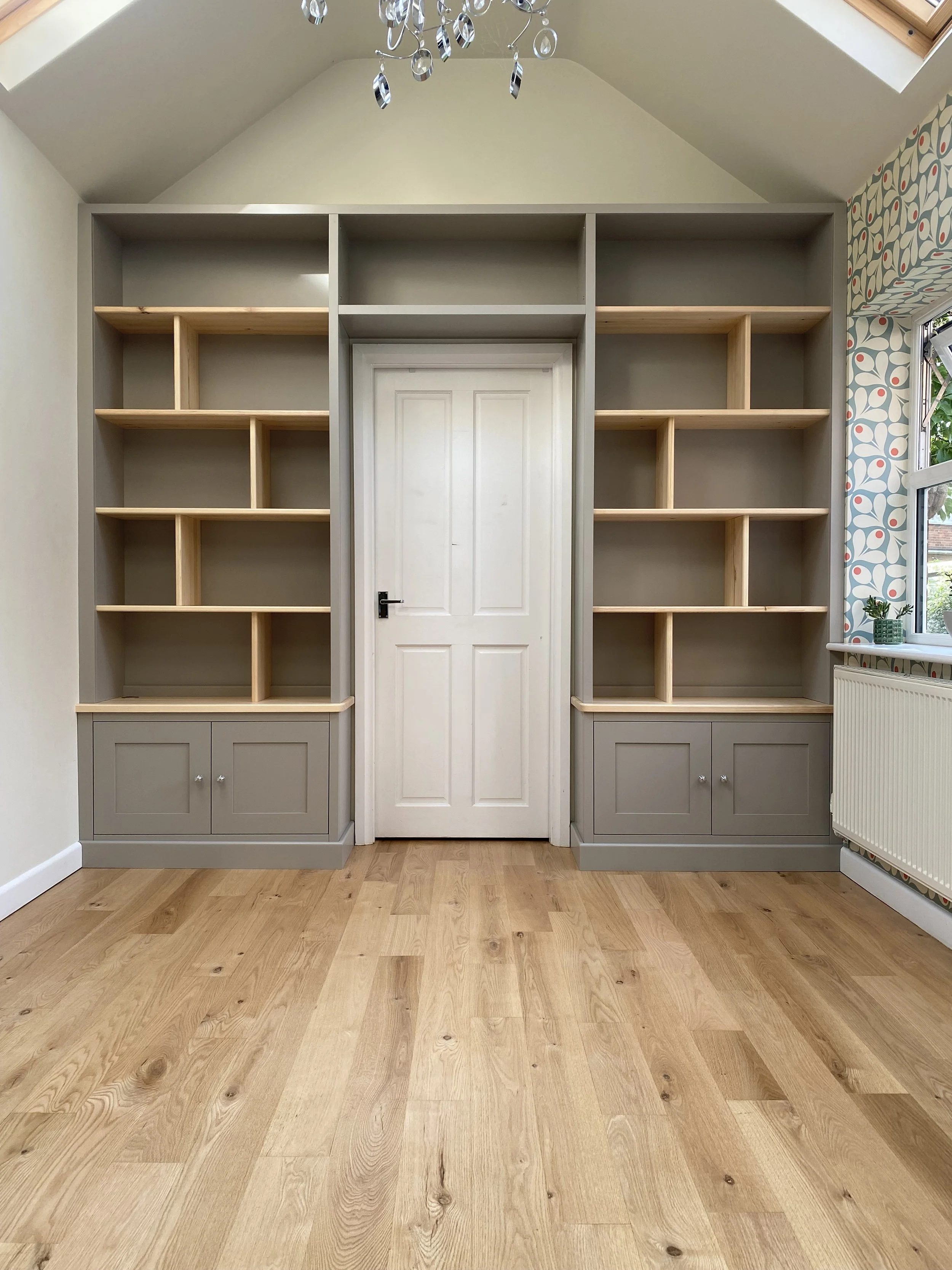Empty built-in shelves with a closed door in the center, in a room with wooden flooring and patterned wallpaper on the right wall.