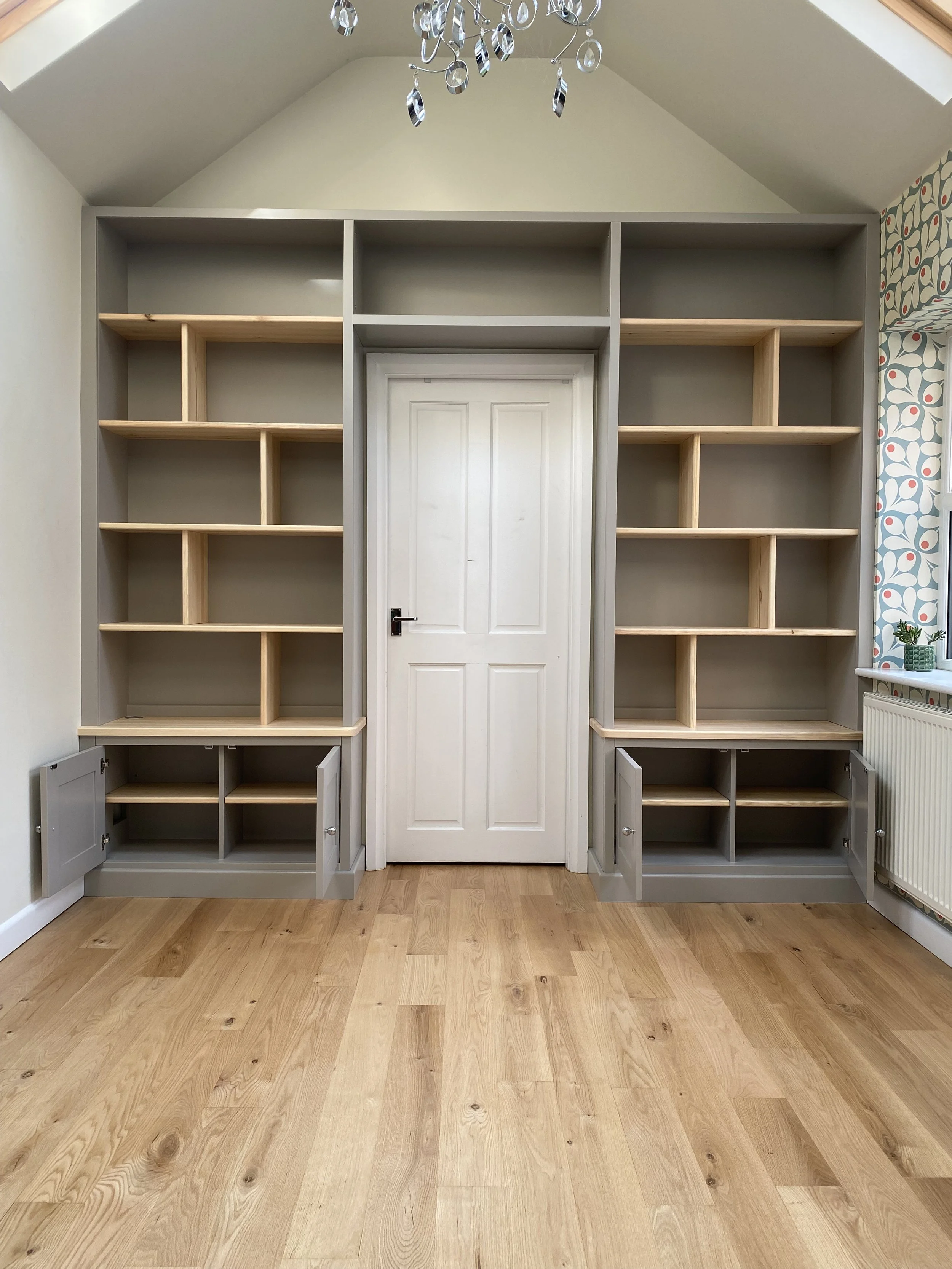 Empty built-in bookshelf with open cabinet doors at the bottom, white door in the center, and patterned wallpaper on the right side, in a room with wooden flooring and a chandelier overhead.
