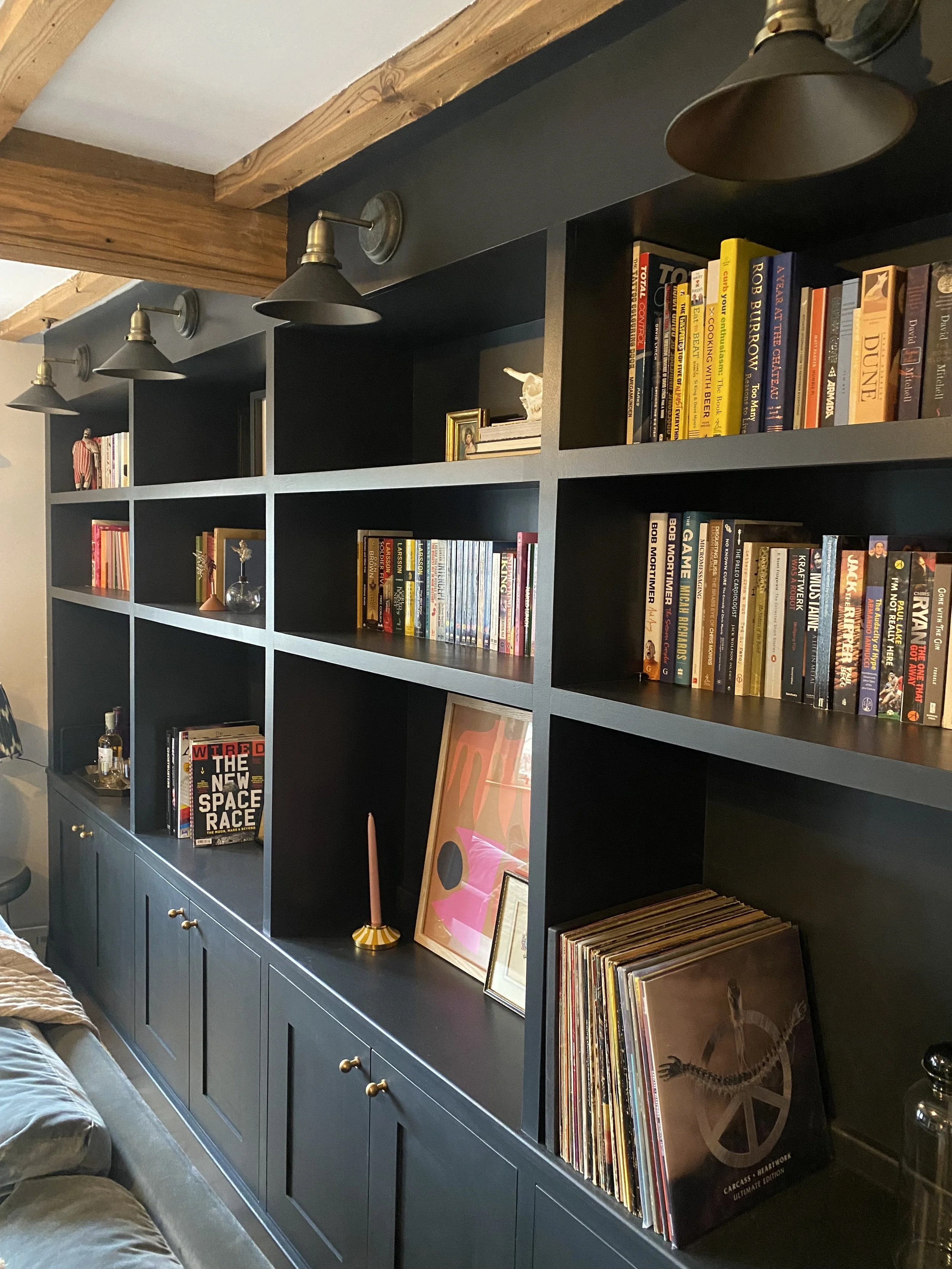 Dark blue bookshelf with multiple compartments containing books, framed pictures, and decorative items, mounted on a wall with exposed wooden beams and fitted with brass hardware.