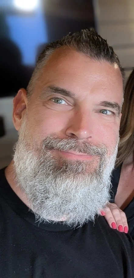 Close-up of a smiling middle-aged man with a beard and short dark hair, wearing a black shirt.