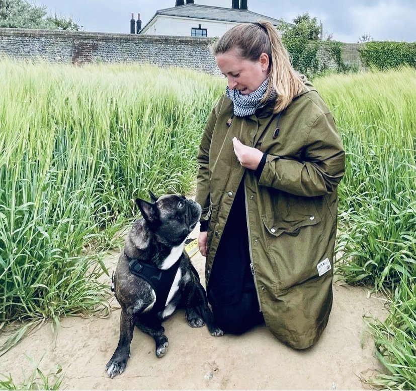 A woman kneeling on a dirt path in a green field with a black and white dog looking up at her. She is wearing a green jacket and a gray scarf, with a partly cloudy sky above.