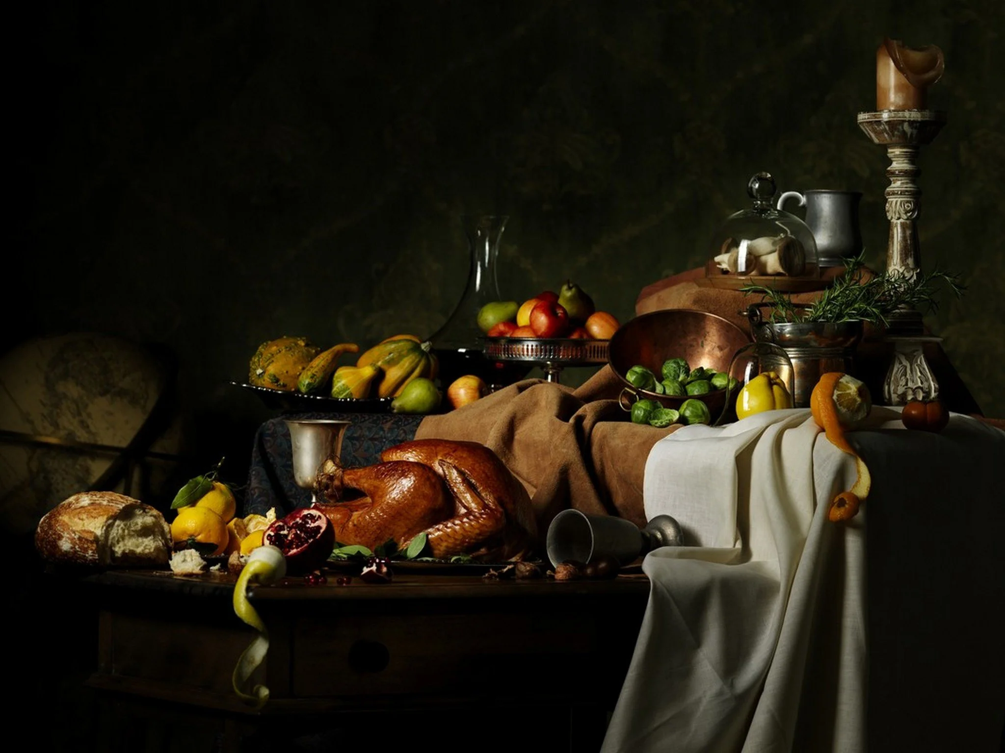 Dark still-life of a Thanksgiving-style feast with a cooked turkey, assorted fruits, vegetables, bread, decorative gourds, a silver goblet, and an ornate candle holder.