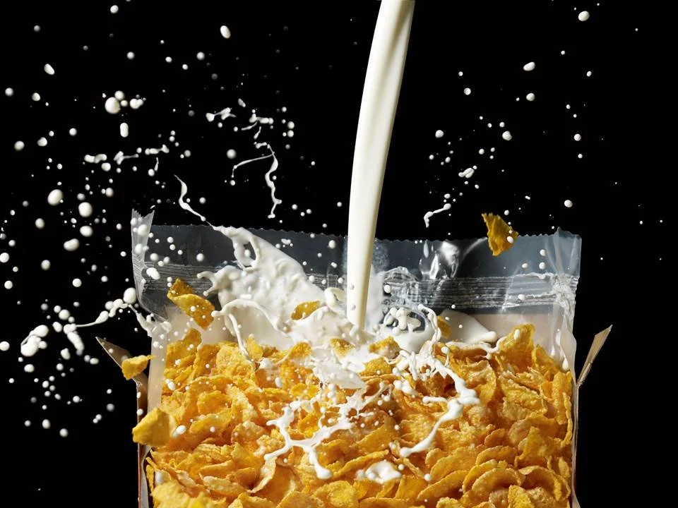 Cut in Half by Food Stylist Charlotte Omnes & Photographer Beth Galton-Milk splashing onto cornflakes in an open cereal bag against a black background.