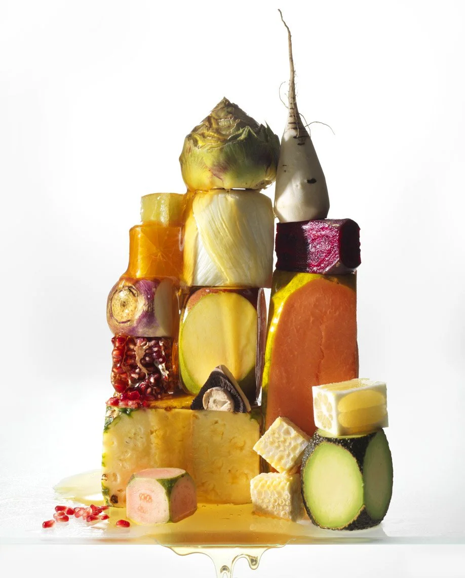 Food Stylist Charlotte Omnes & Photographer Francine Zaslow-Assorted fruits and vegetables stacked artistically, including pineapple, artichoke, zucchini, sweet potato, beetroot, and honeycomb, with scattered pomegranate seeds and dripping honey.