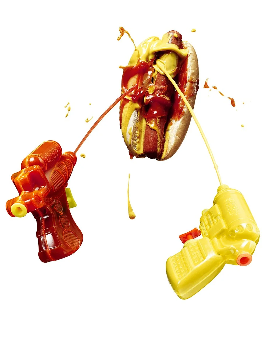 A hot dog with ketchup and mustard being squirted onto it from toy water guns.
